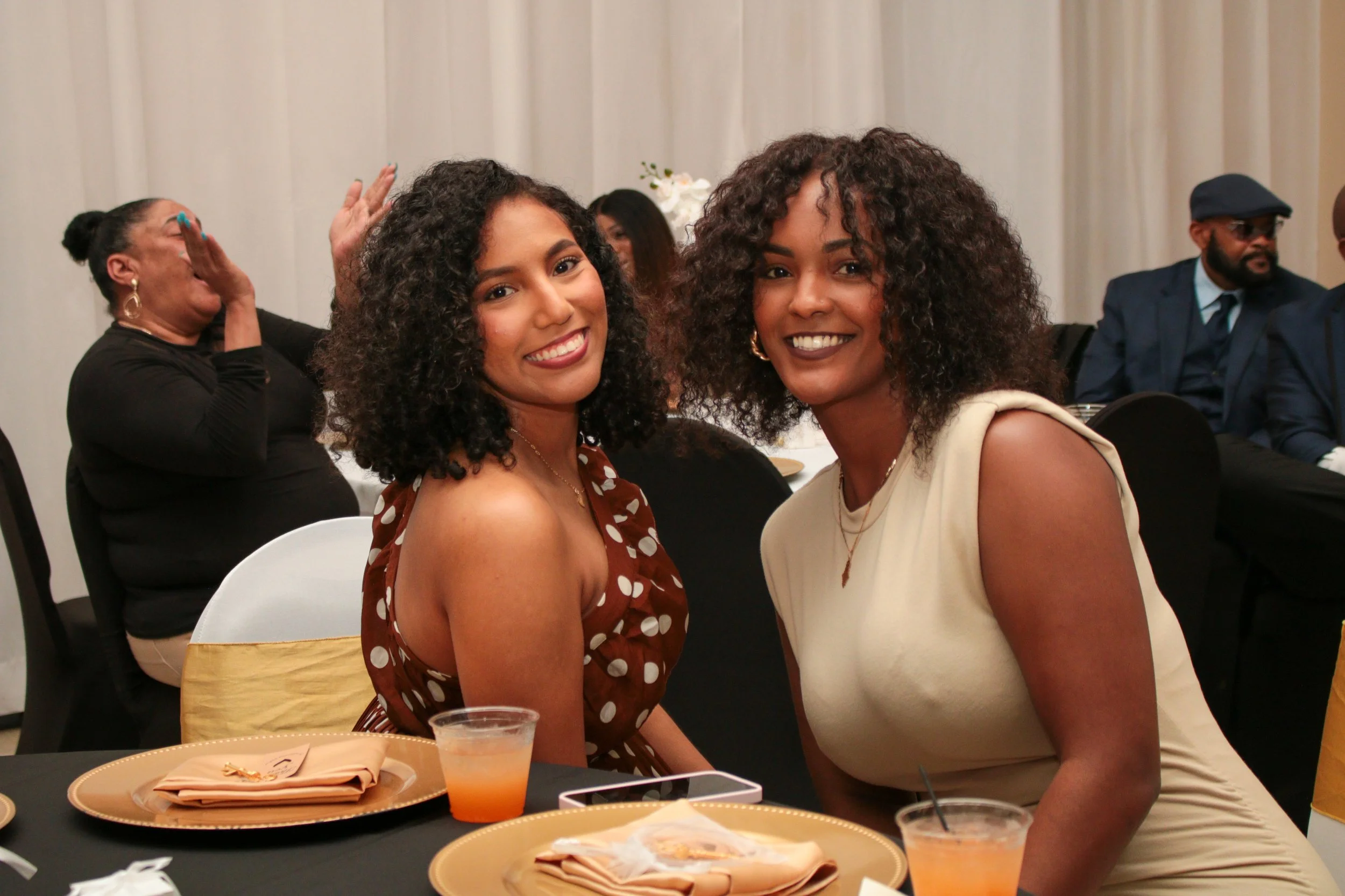 Two women sitting at a banquet table, smiling at the camera, with others in the background at the event.