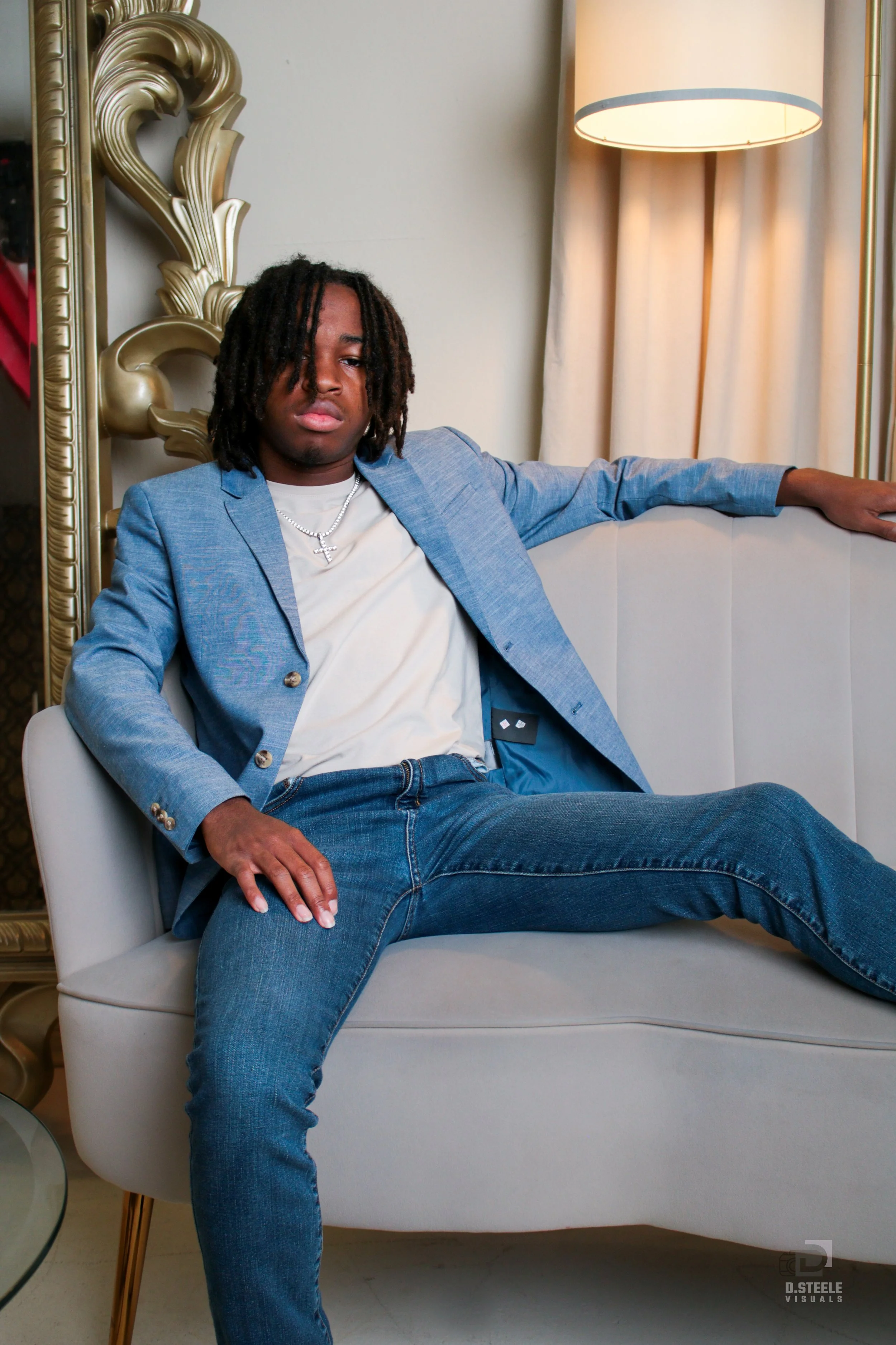 A young man with dreadlocks wearing a light blue blazer, white t-shirt, and jeans, sitting on a cream-colored sofa with one arm resting on the back and one hand on his thigh, in a room with a beige curtain and a table lamp.
