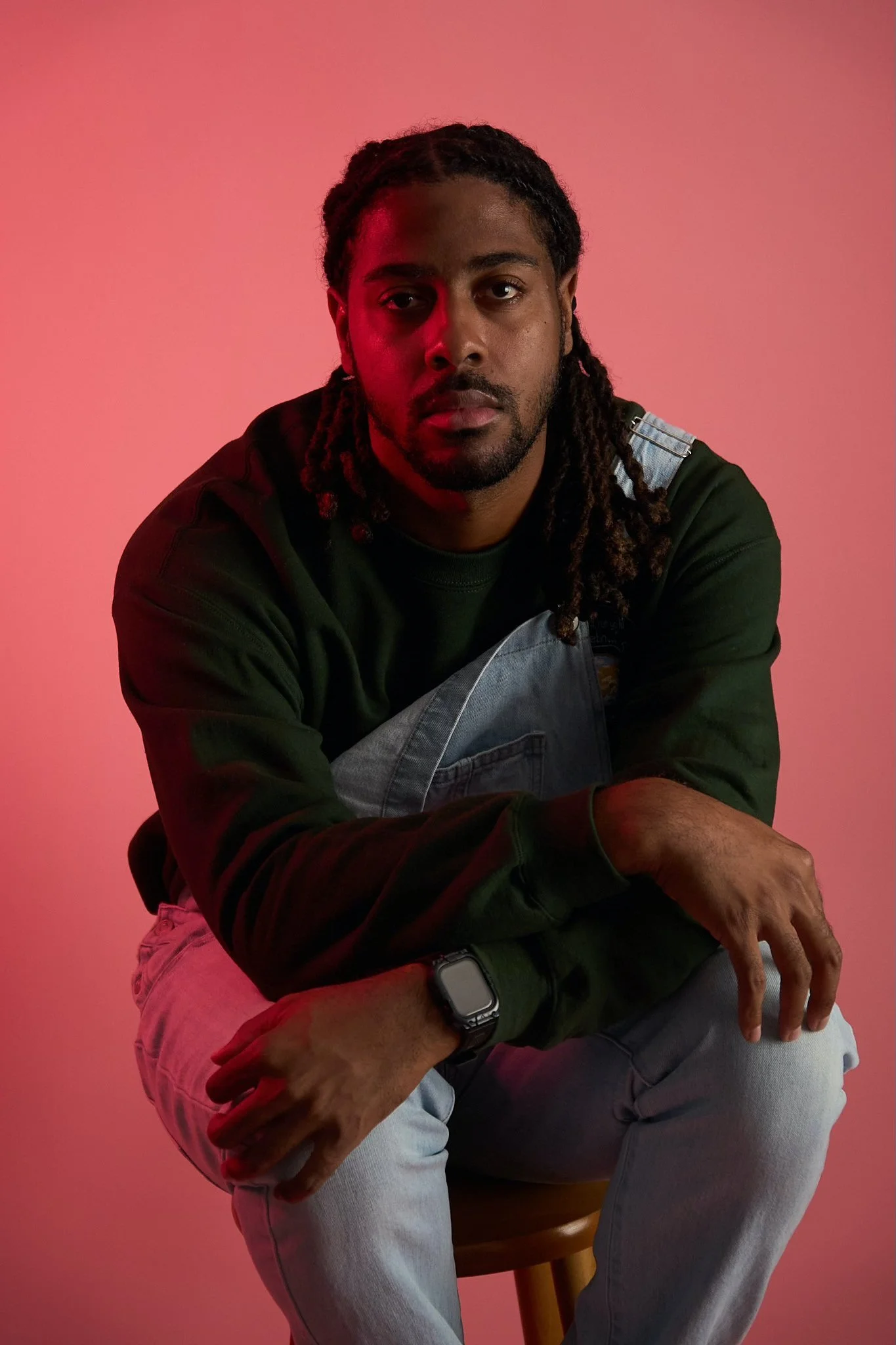 A man with dreadlocks wears a green sweatshirt and light blue jeans, sitting on a wooden stool against a pink background, with a smartwatch on his left wrist.