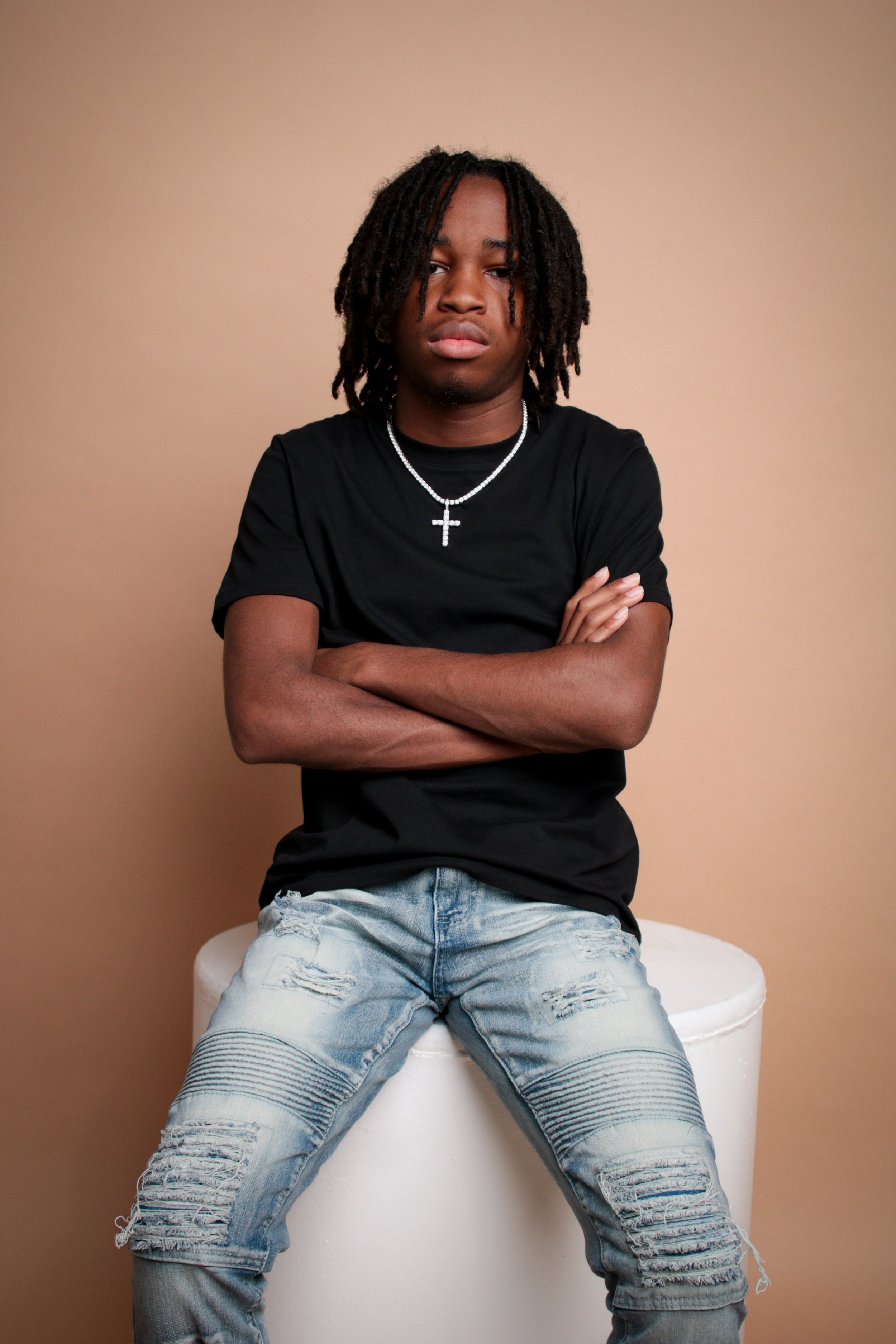 A young man with dreadlocks wearing a black T-shirt, ripped jeans, and a cross necklace, sitting with arms crossed against a beige background.