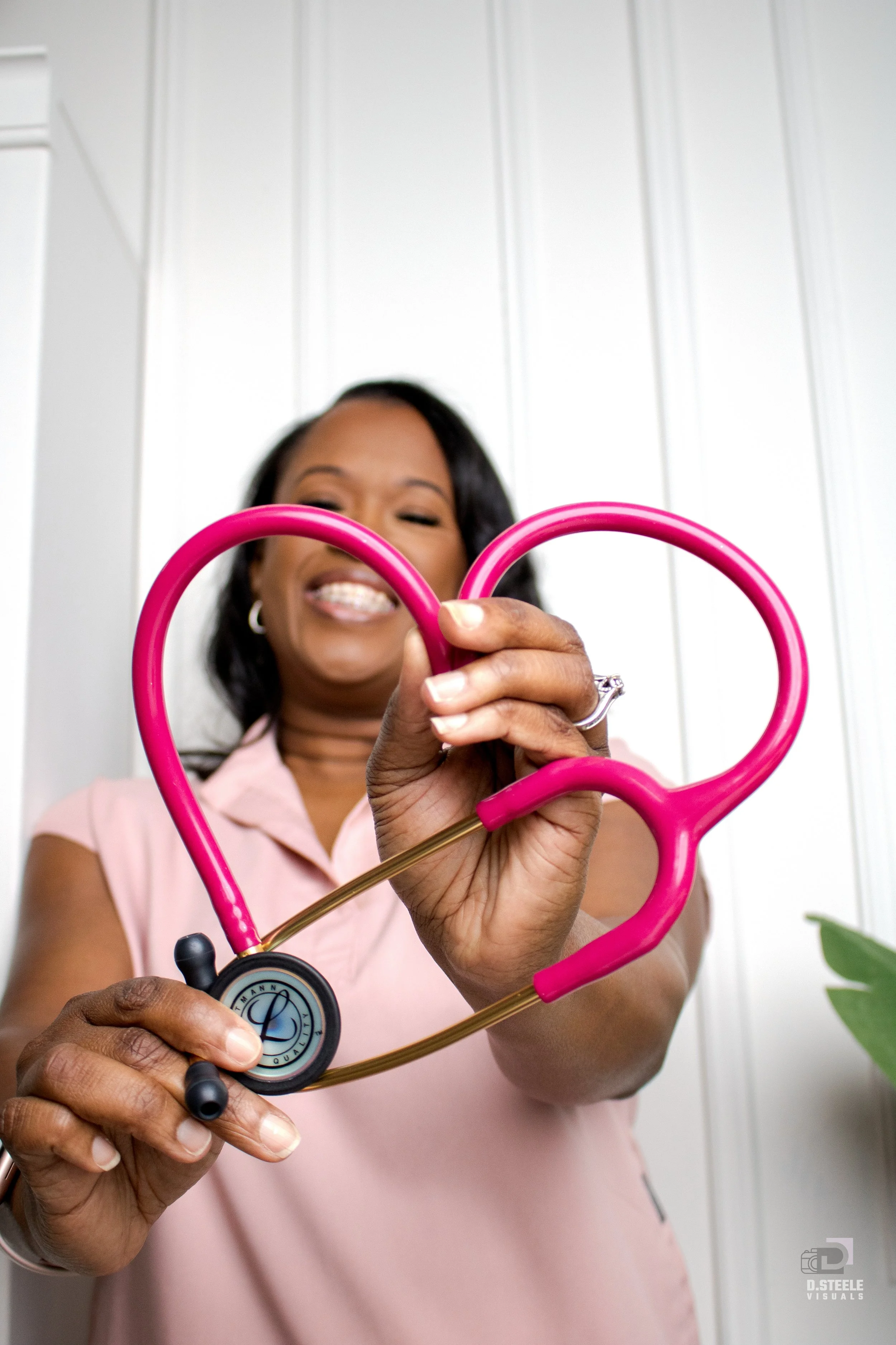 A woman holding a pink heart-shaped stethoscope, smiling, with a white background.