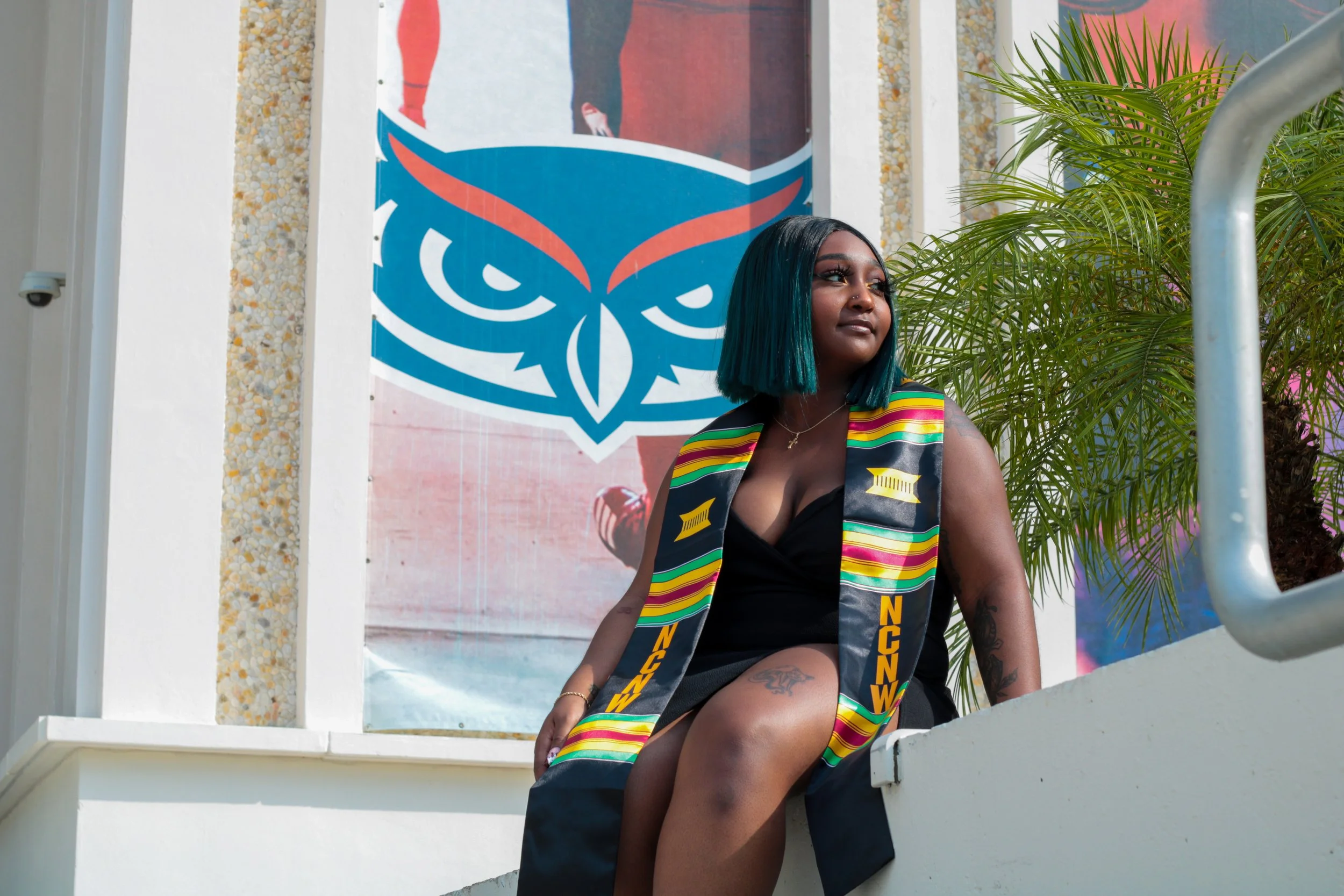 A woman with university graduation attire sitting on a ledge outdoors, with a large owl logo on the wall behind her and green palm leaves nearby.