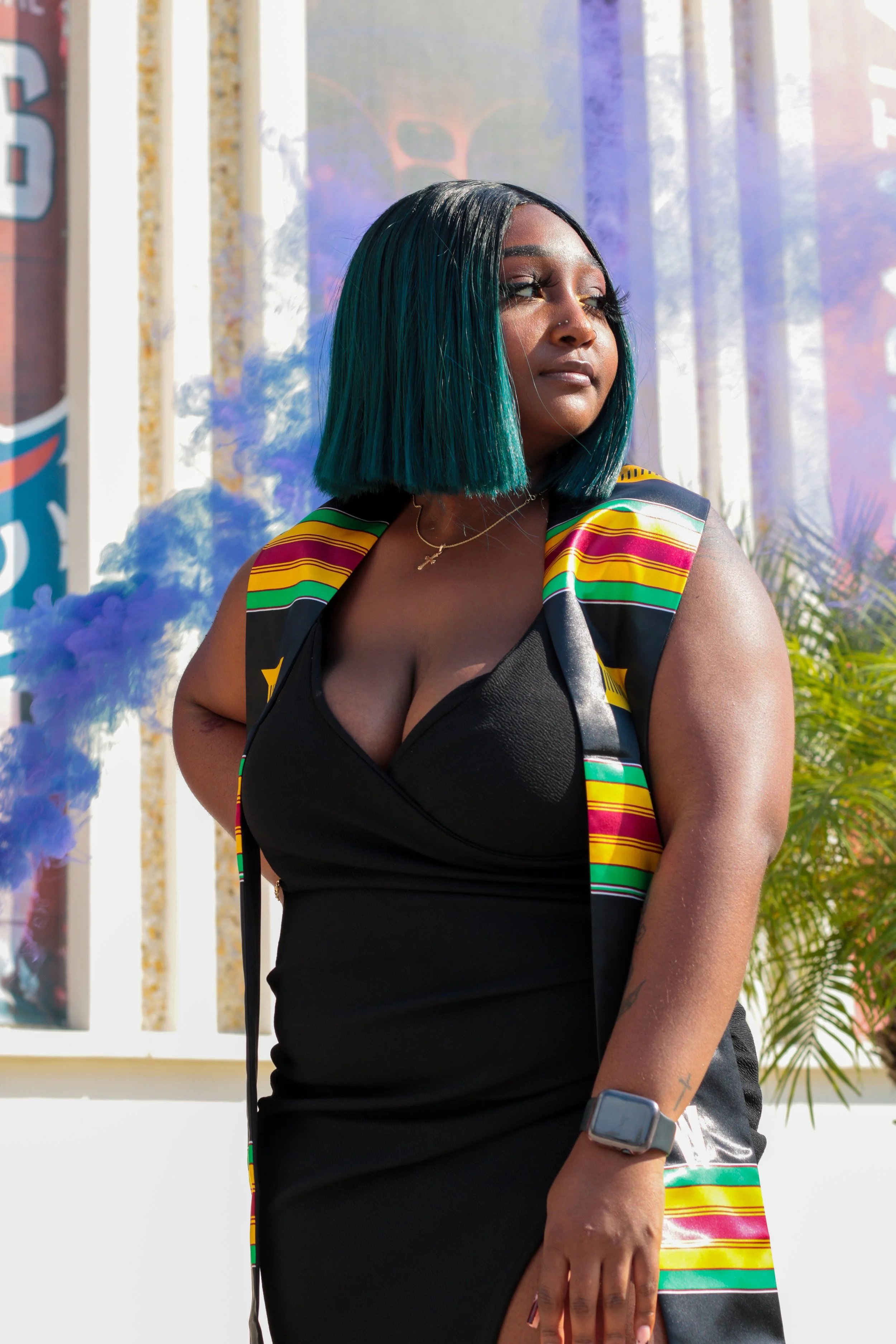A woman with shoulder-length, teal-colored hair stands outdoors in sunlight, wearing a black dress with a colorful striped vest, a smartwatch, and a necklace. She is looking to the side with a serious expression.