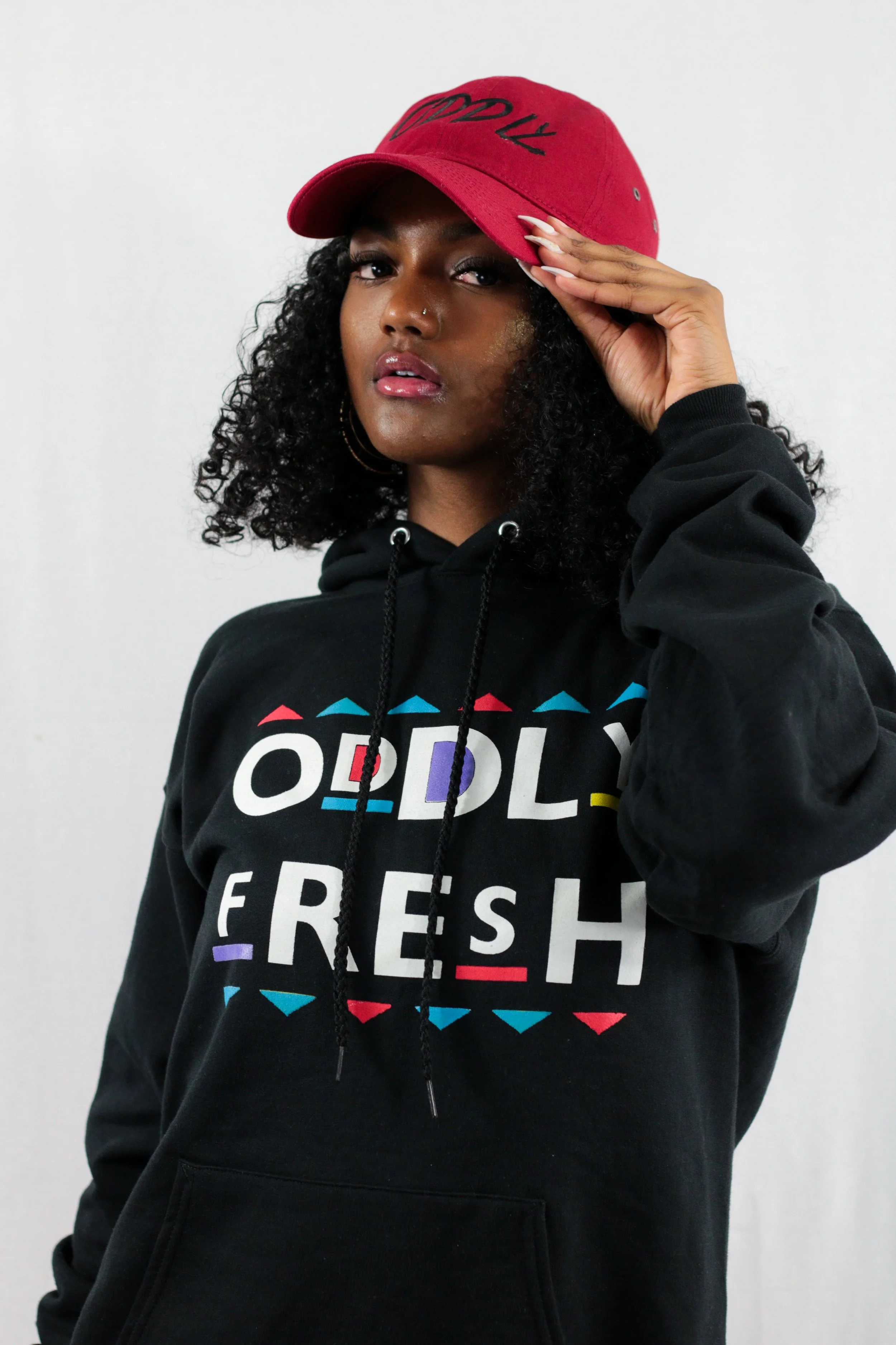 A woman wearing a red baseball cap and black hoodie with colorful text and graphics, posing against a plain white background.