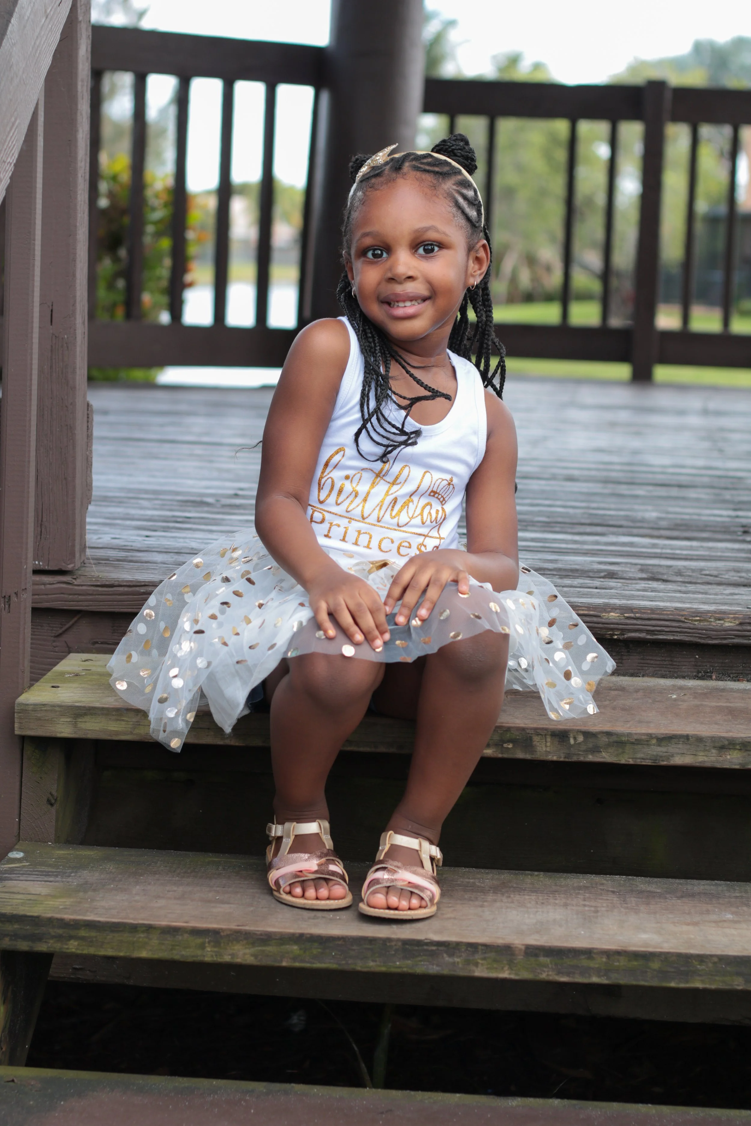 A young girl sitting on steps, wearing a white dress with gold accents and sandals, smiling outdoors with a wooden deck and green trees in the background.