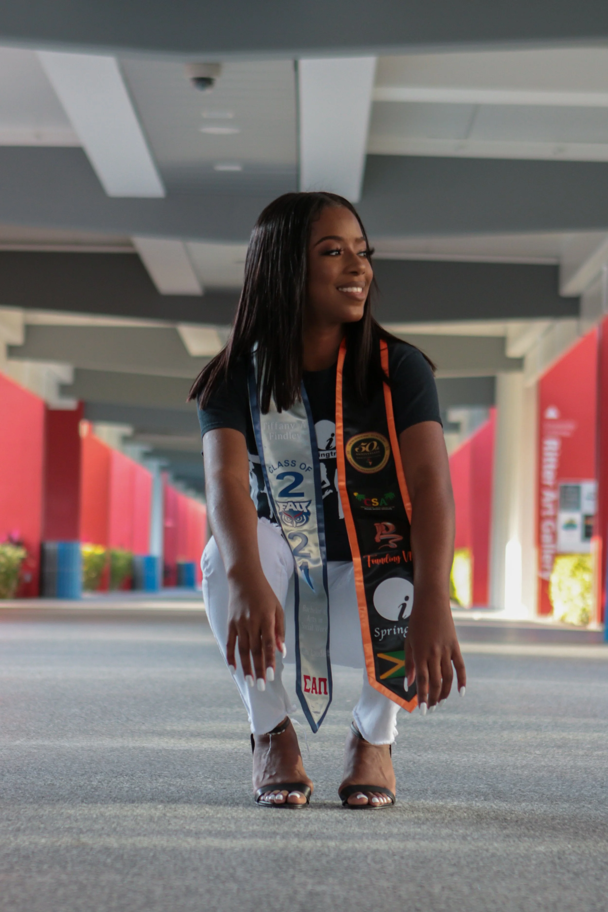 Woman in black shirt and white pants crouching under a bridge at a graduation event, with honorary sashes and medals.