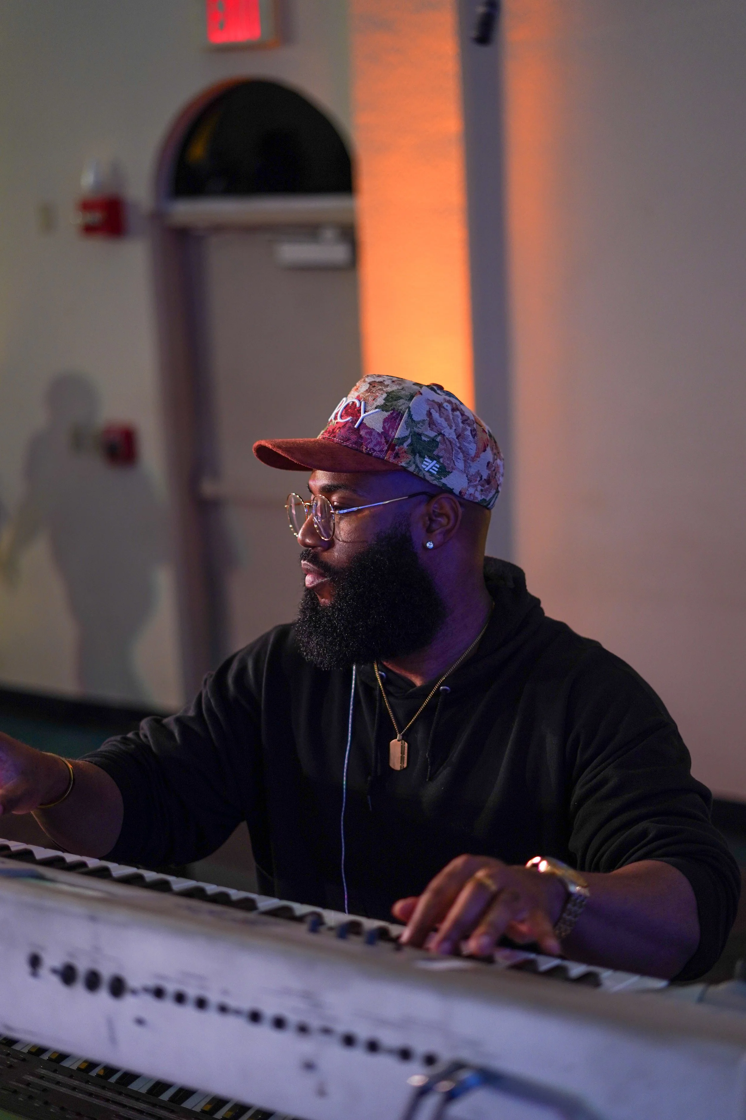 A man with a beard and glasses playing the piano, wearing a floral baseball cap, a black hoodie, and jewelry, in a dimly lit room with colorful lighting.