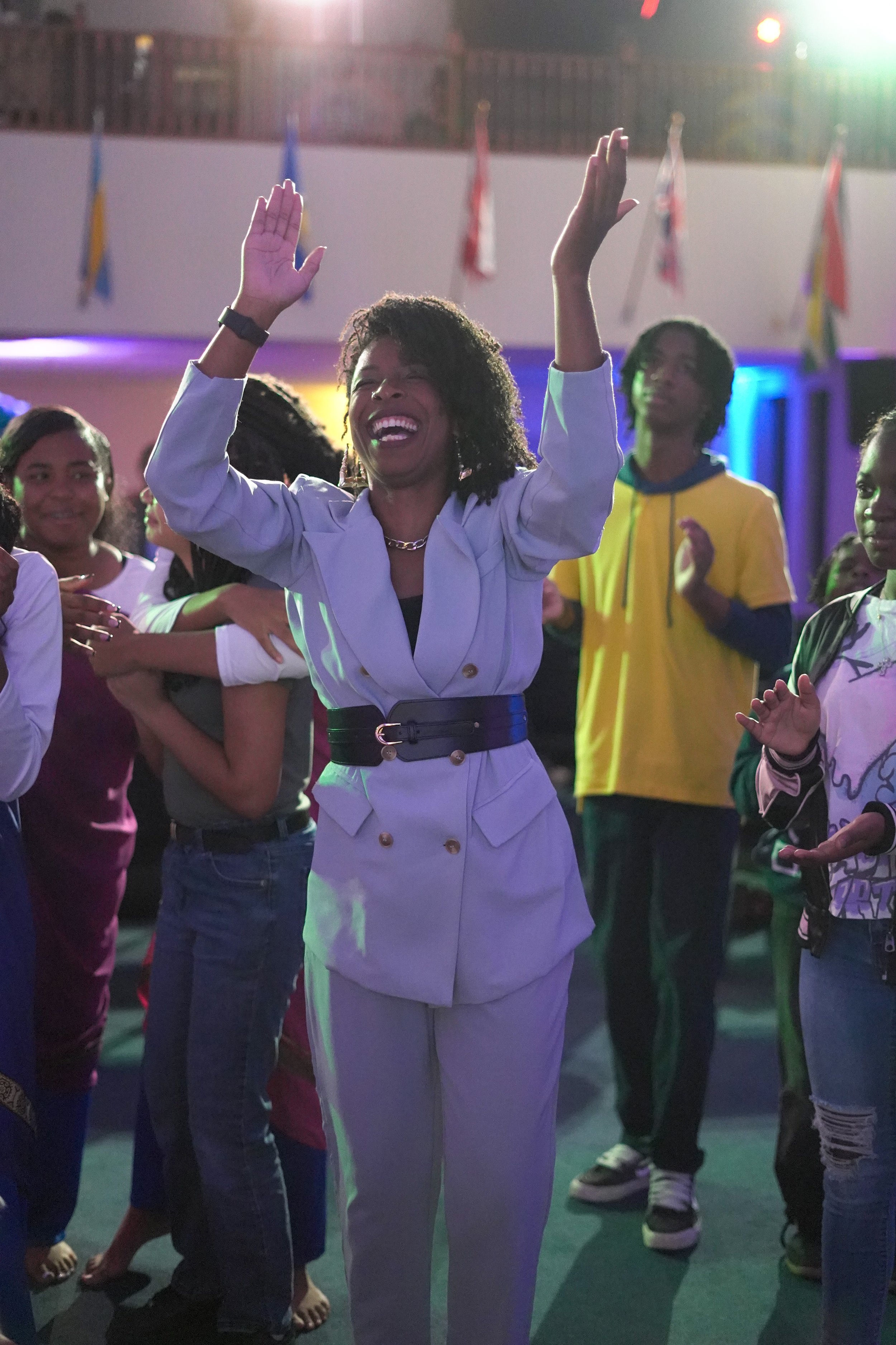 A joyful woman in a white suit with a black belt, raising her arms and smiling at a lively party or celebration surrounded by diverse people.