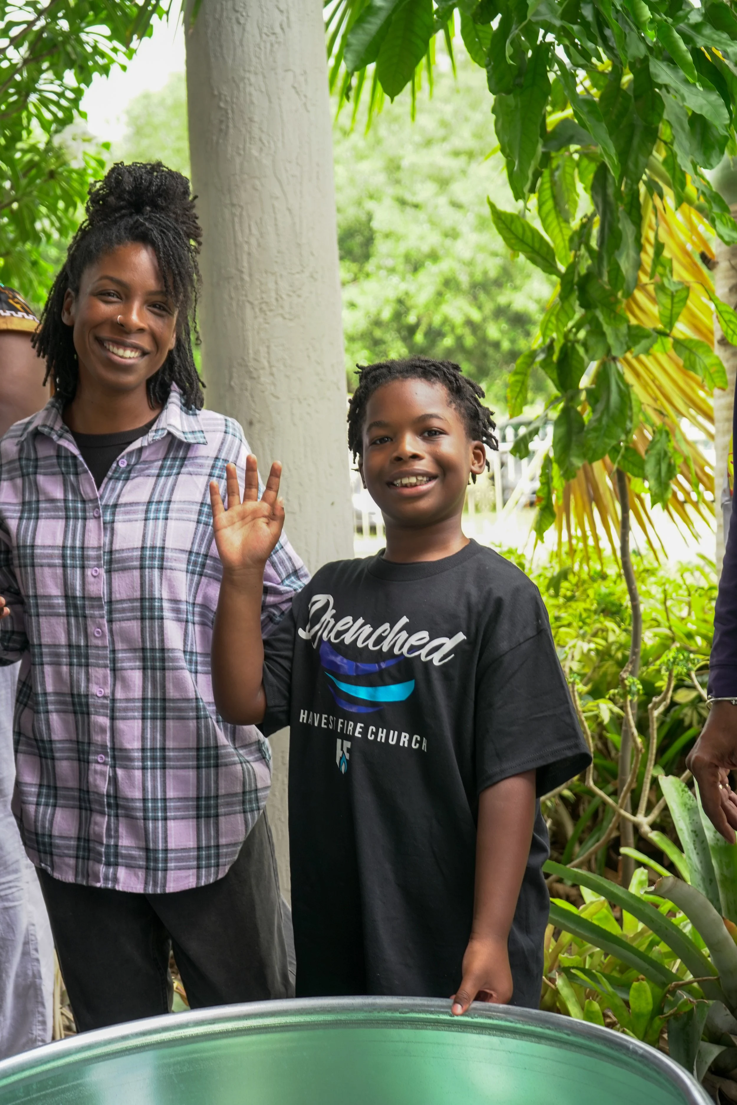 A woman and a boy standing outdoors, smiling and waving at the camera, surrounded by lush green plants and trees.