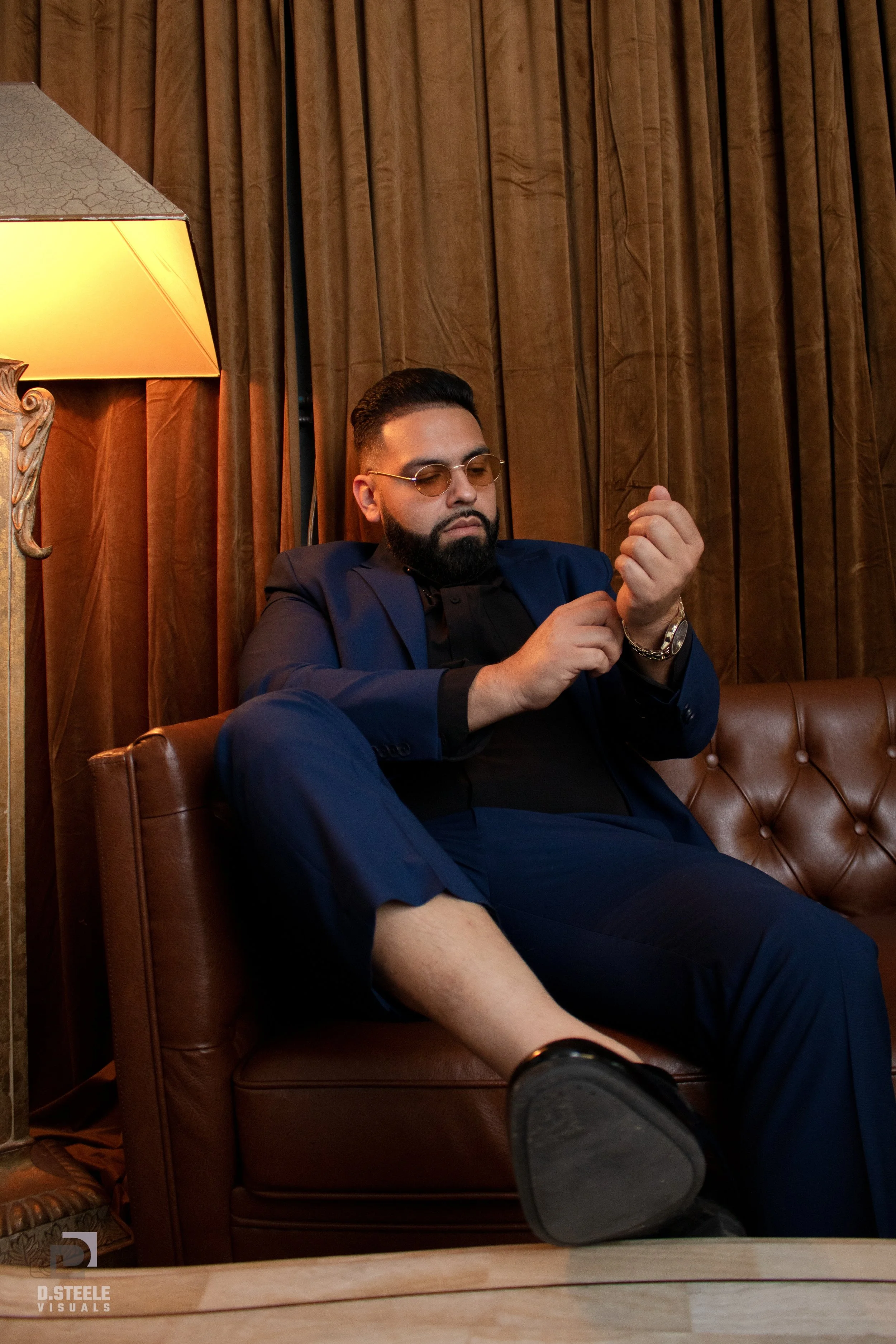 A man with dark hair and beard, wearing glasses, a blue suit, and a black shirt, sits on a brown leather couch in a dimly lit room with brown curtains. He is looking at his watch, with a relaxed posture, one leg crossed over the other.