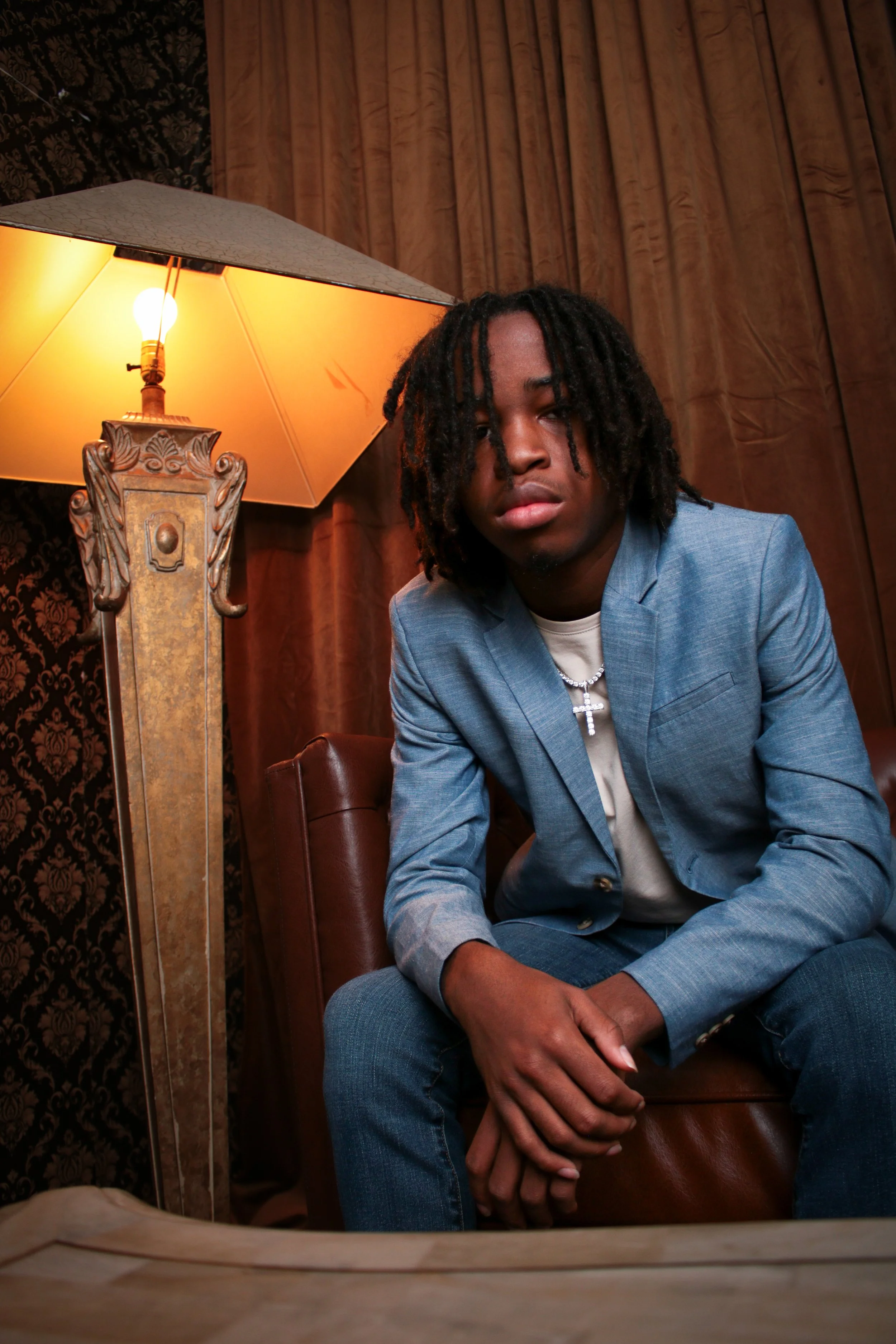 A young man with Dreadlocks wearing a light blue blazer, white shirt, and necklace with a cross, sitting on a leather chair in front of a vintage lamp with warm lighting.