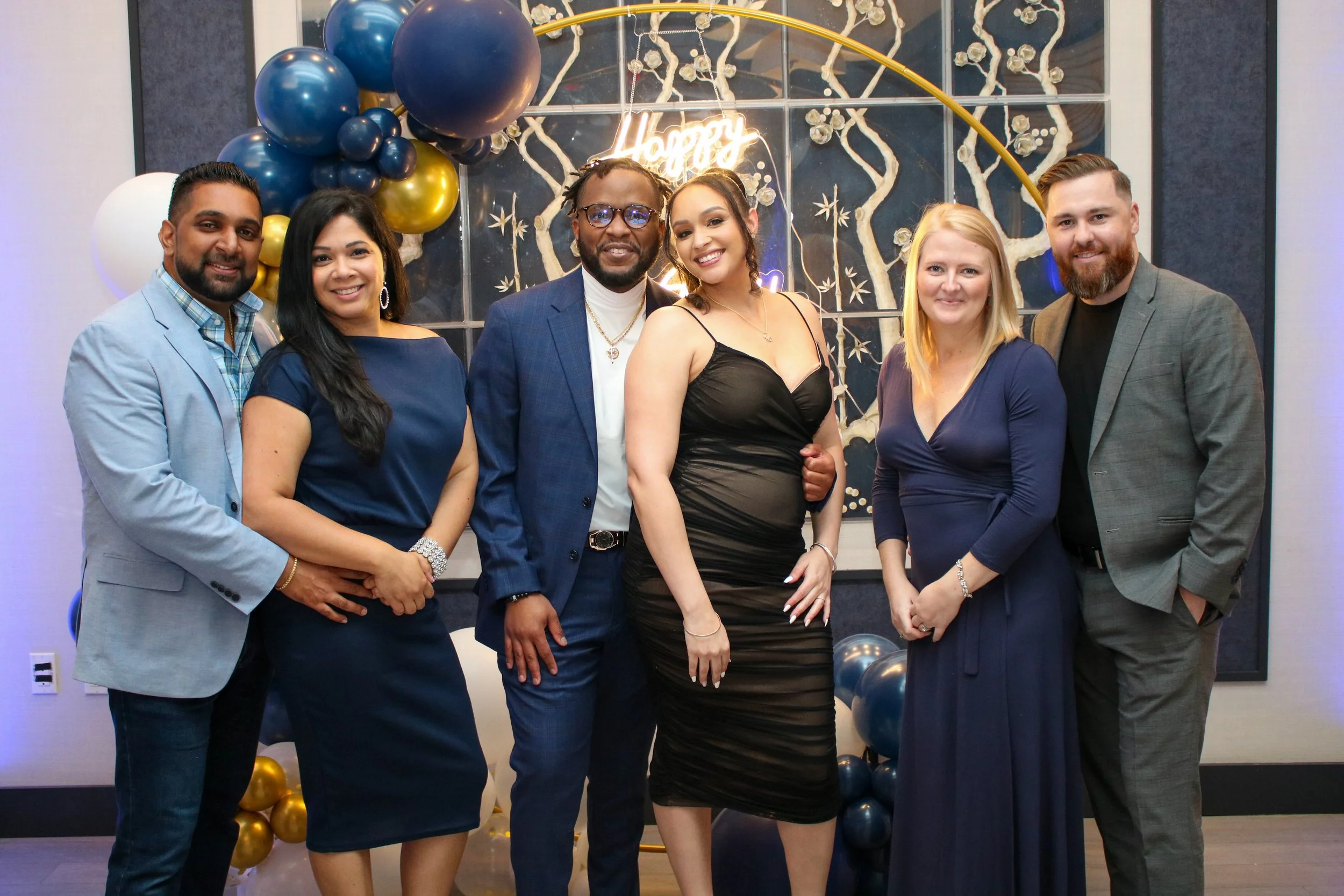 A group of seven people dressed in formal attire, standing together at a celebration with balloons and a decorative backdrop, smiling for the photo.