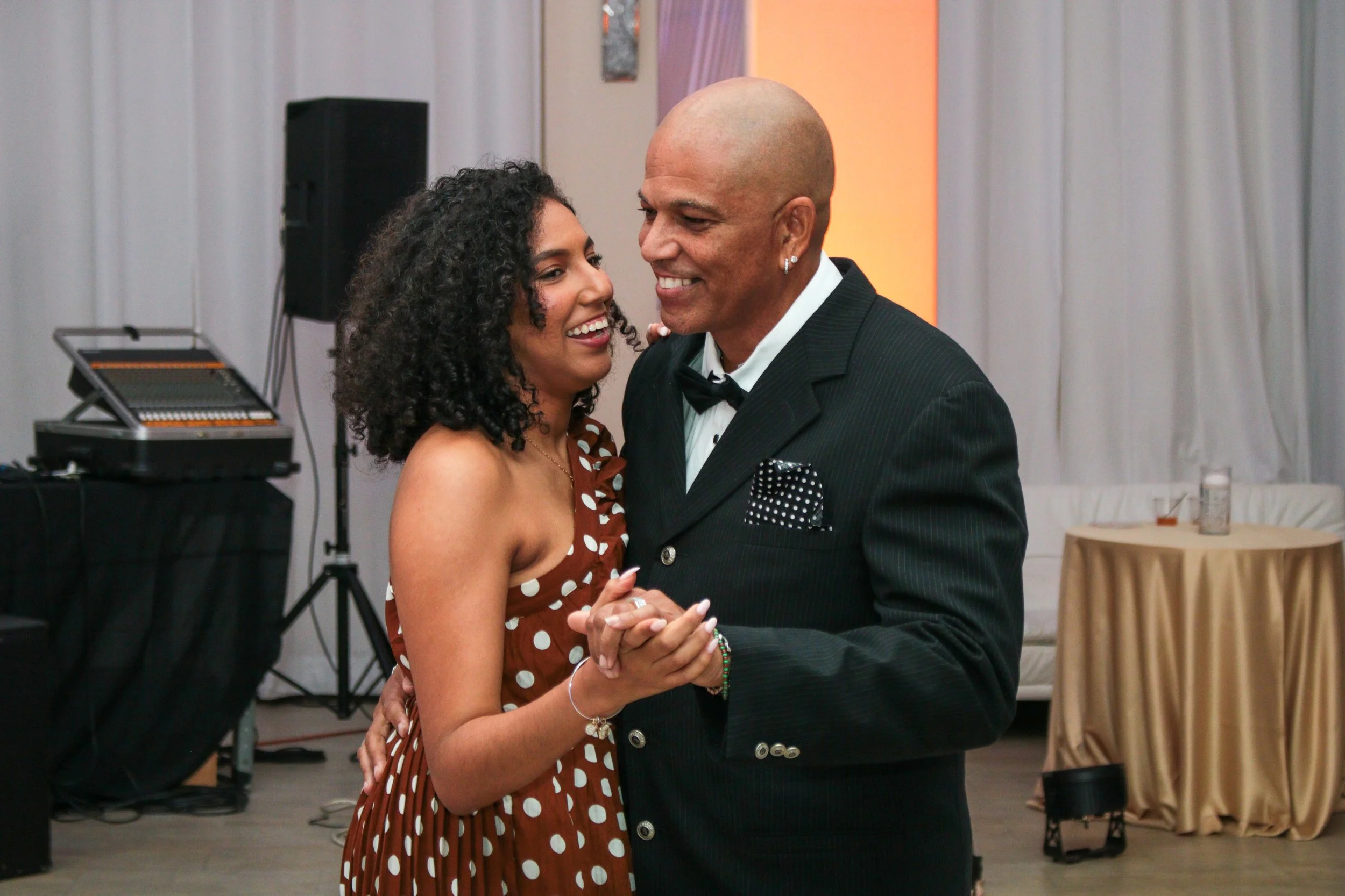 A couple dancing at a formal event, the woman with curly hair in a brown polka dot dress and the man in a black tuxedo with a pocket square, smiling and holding hands.