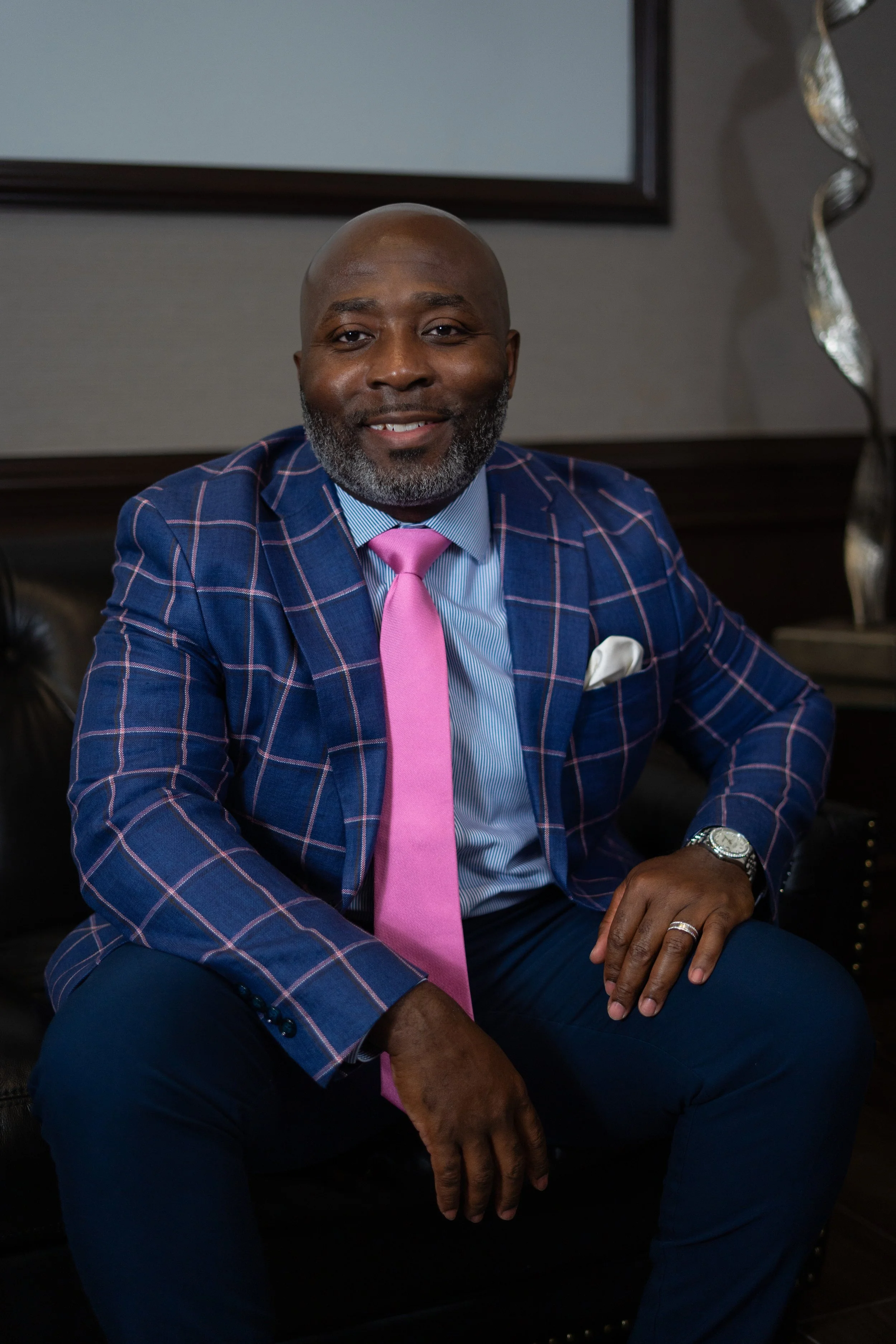 A man wearing a blue checkered suit, pink tie, and light blue shirt, sitting on a leather sofa, smiling at the camera.