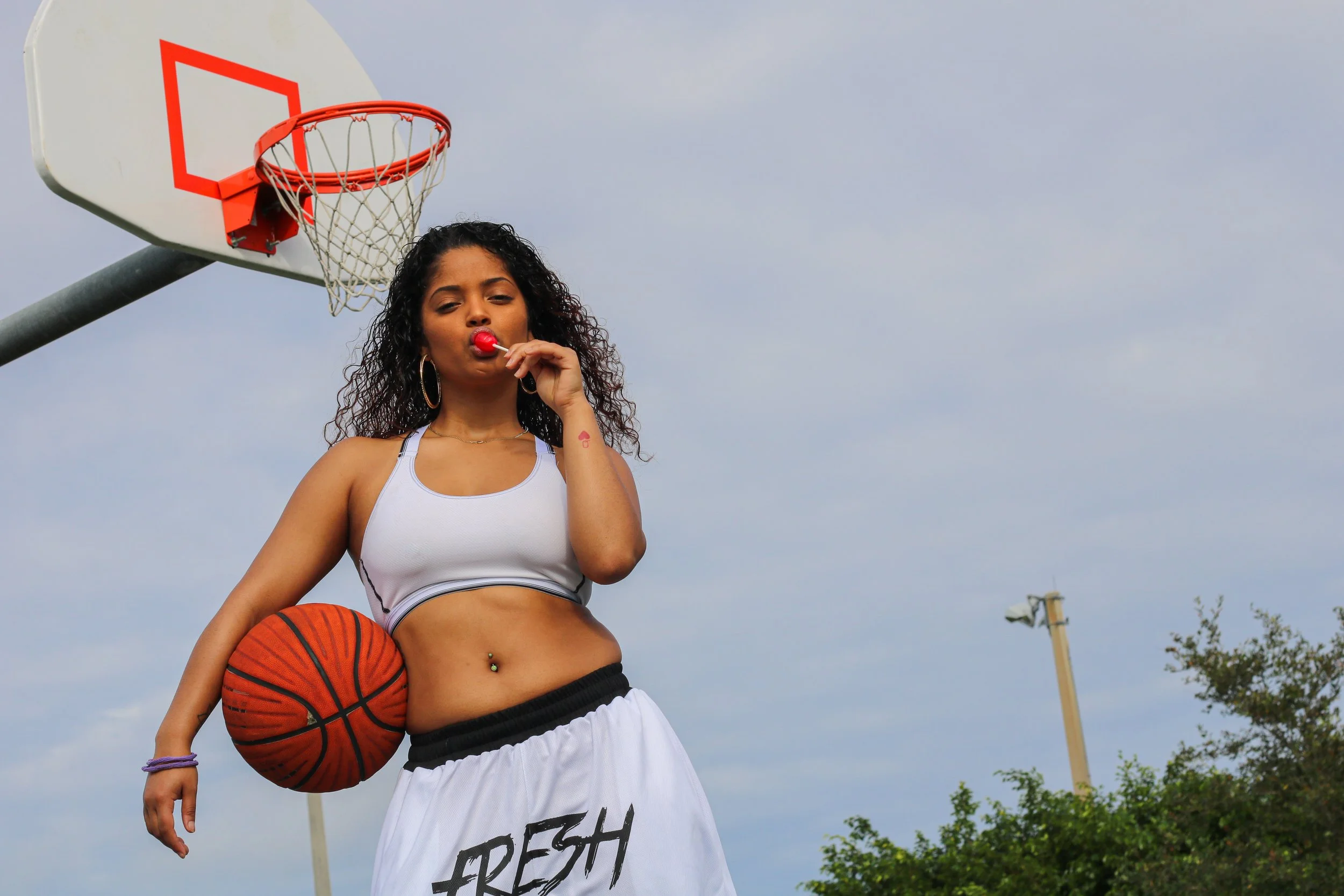 Young woman in white sports bra and shorts holding a basketball under her arm, standing under a basketball hoop outdoors, blowing a red lollipop, with a cloudy sky and some greenery in the background.