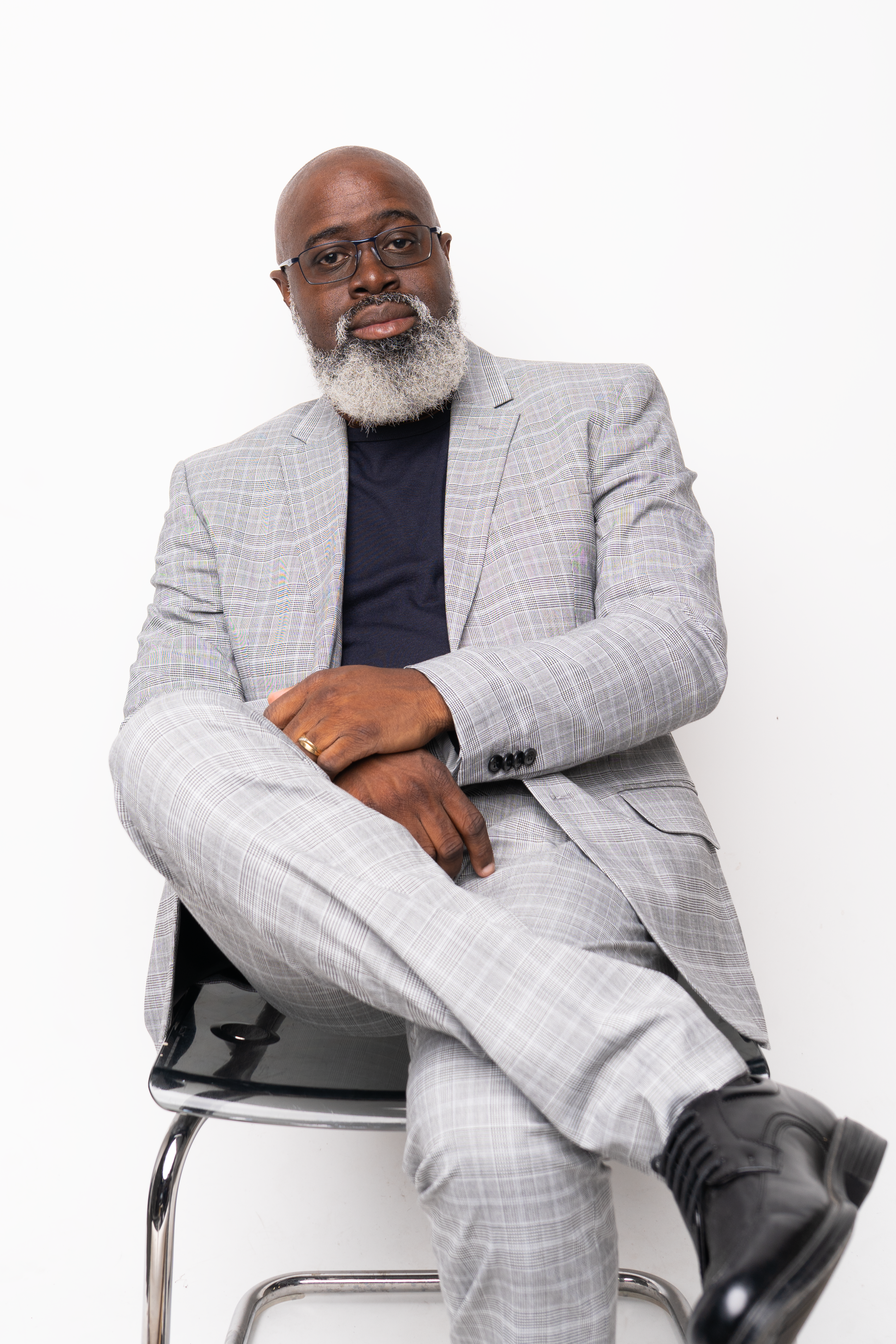 A mature man with a gray beard and glasses sitting on a black chair, wearing a light gray checked suit, black shirt, and black shoes, with a white background.