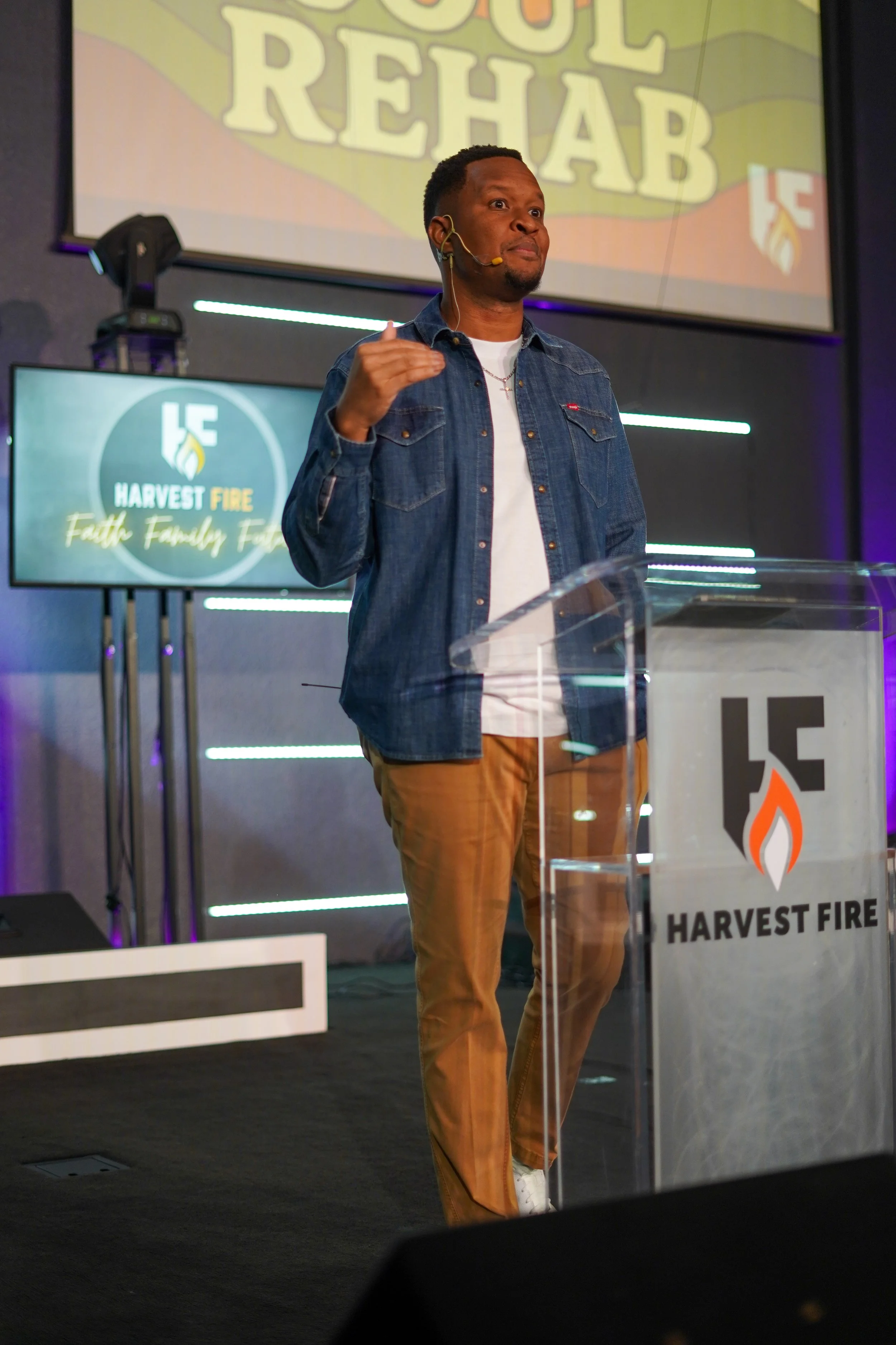 A man standing at a podium with a Harvest Fire logo, speaking into a microphone headset. Behind him are two screens displaying the same logo, and the background features striped lighting.