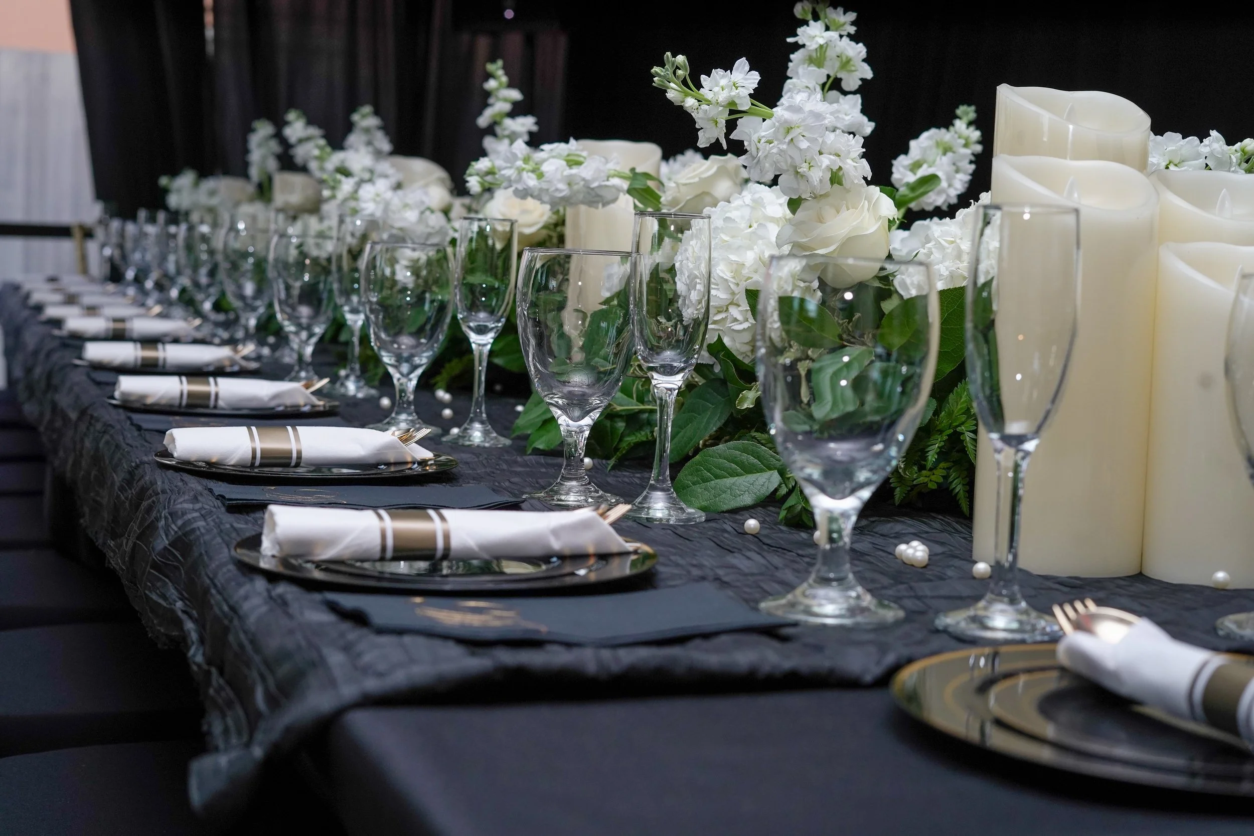 Elegant table setting with black tablecloth, white floral centerpieces, candles, and glassware, prepared for a formal event or wedding reception.