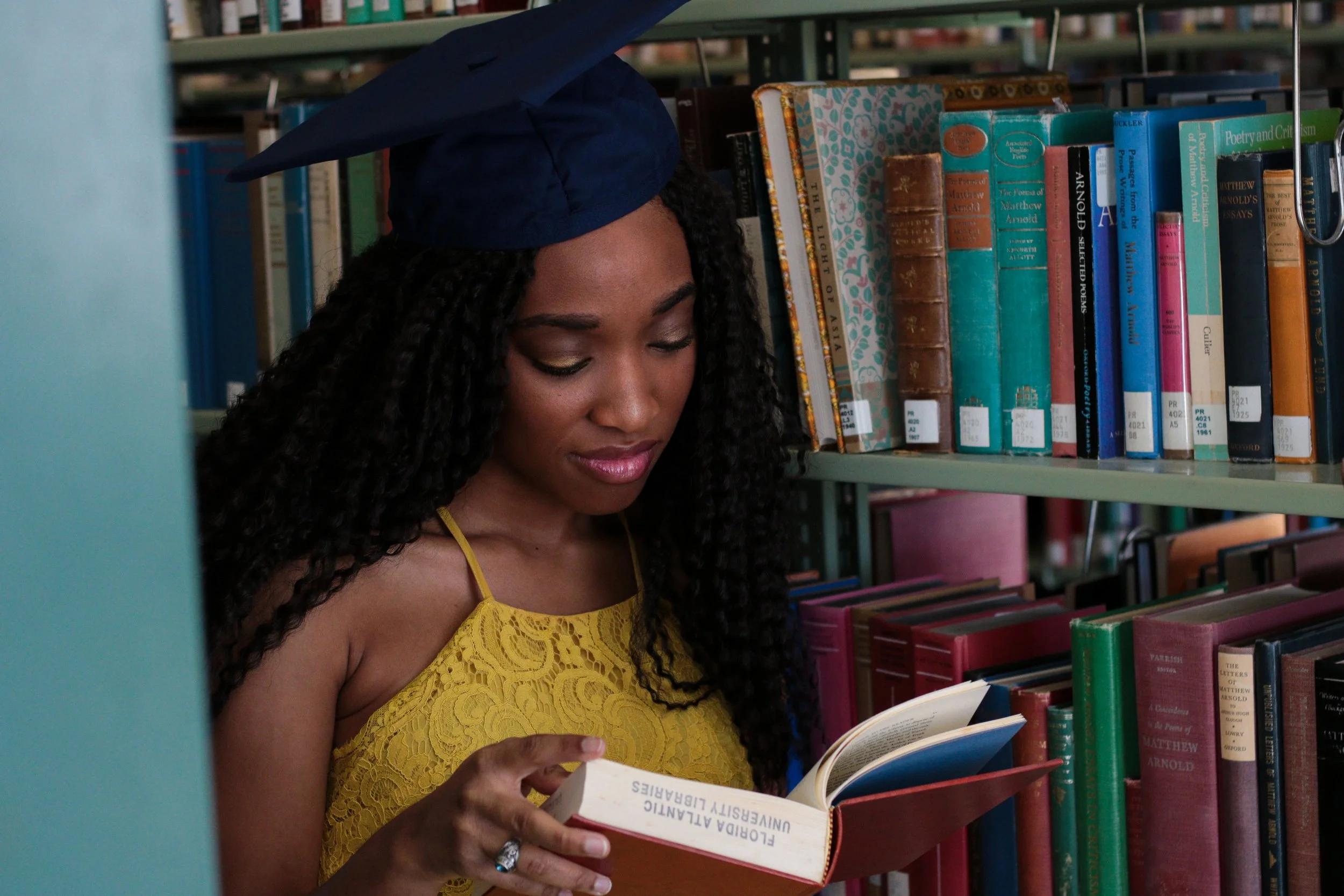 Young woman wearing a blue graduation cap and yellow dress, reading a book titled 'Florida Atlantic University Libraries' in a library aisle filled with colorful books.