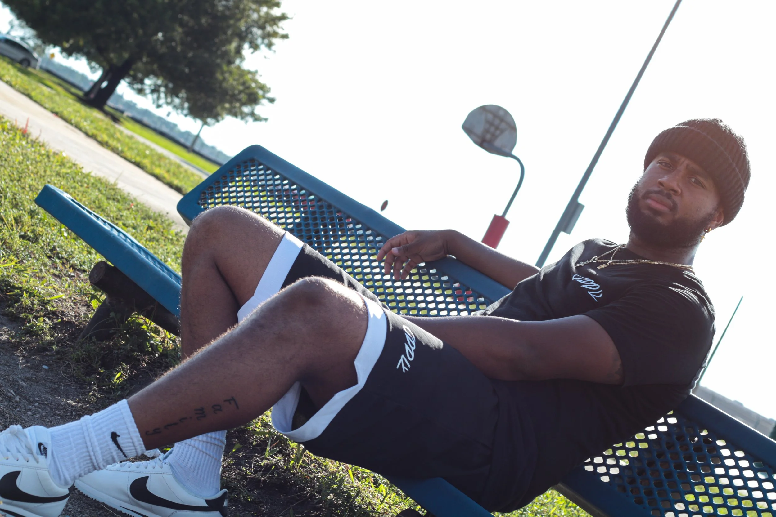 A man with a beard wearing a black beanie, black t-shirt, and black shorts sitting on a blue park bench outdoors on a sunny day.