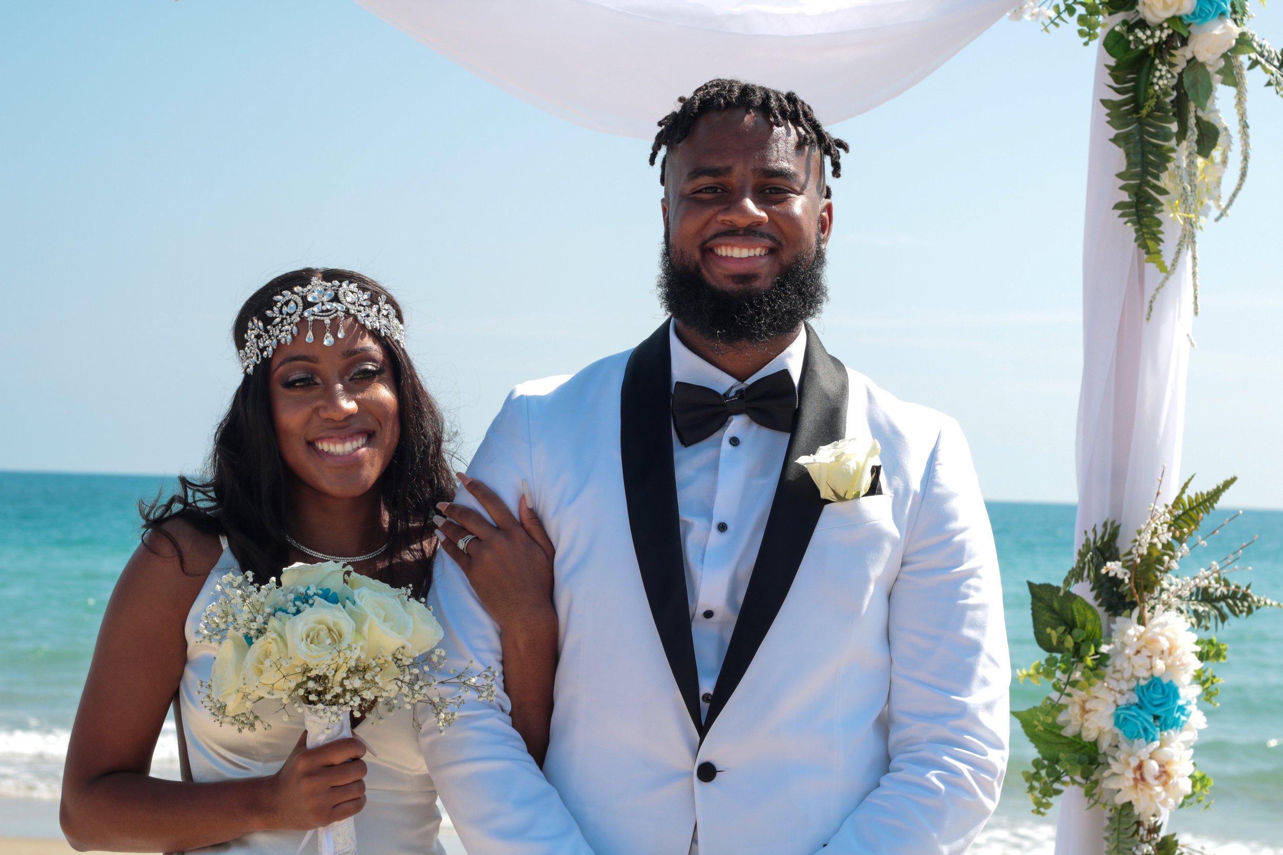 A happy bride and groom posing under a decorated arch on a beach. The bride is holding a bouquet of white roses and wearing a jeweled headband, while the groom is in a white tuxedo with black lapels and a black bow tie, with an ocean and blue sky in the background.