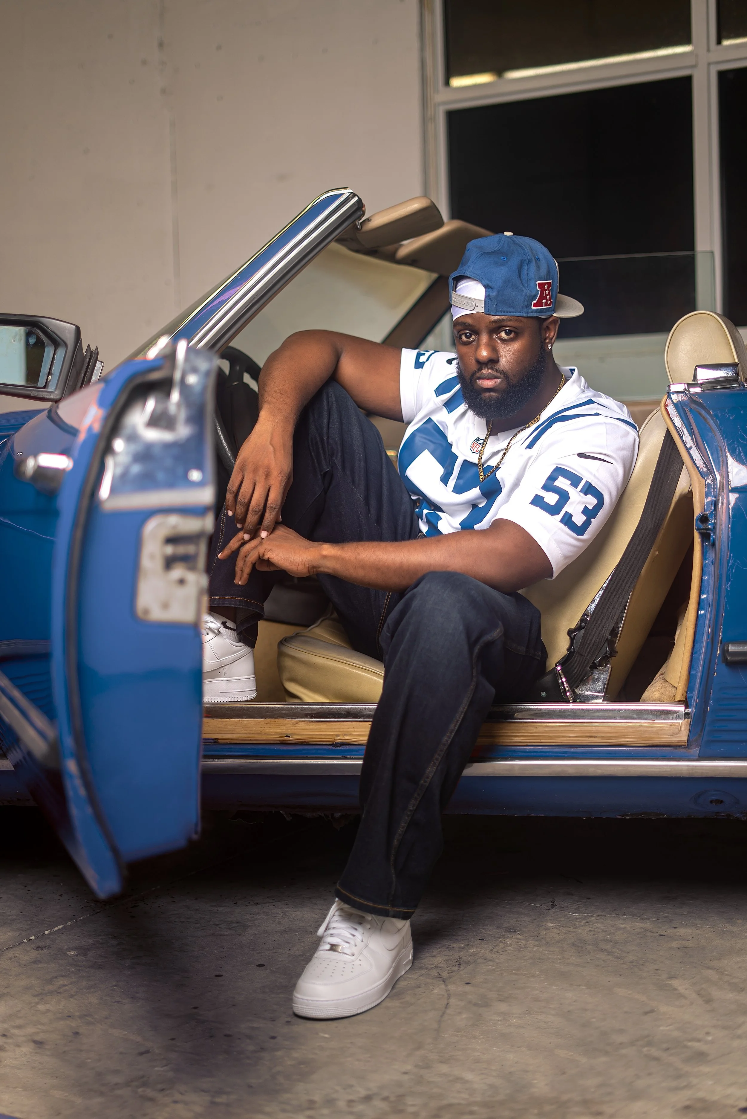 A man is sitting inside a blue vintage convertible car, wearing a white sports jersey, blue jeans, white sneakers, and a blue and grey cap, with a serious expression.
