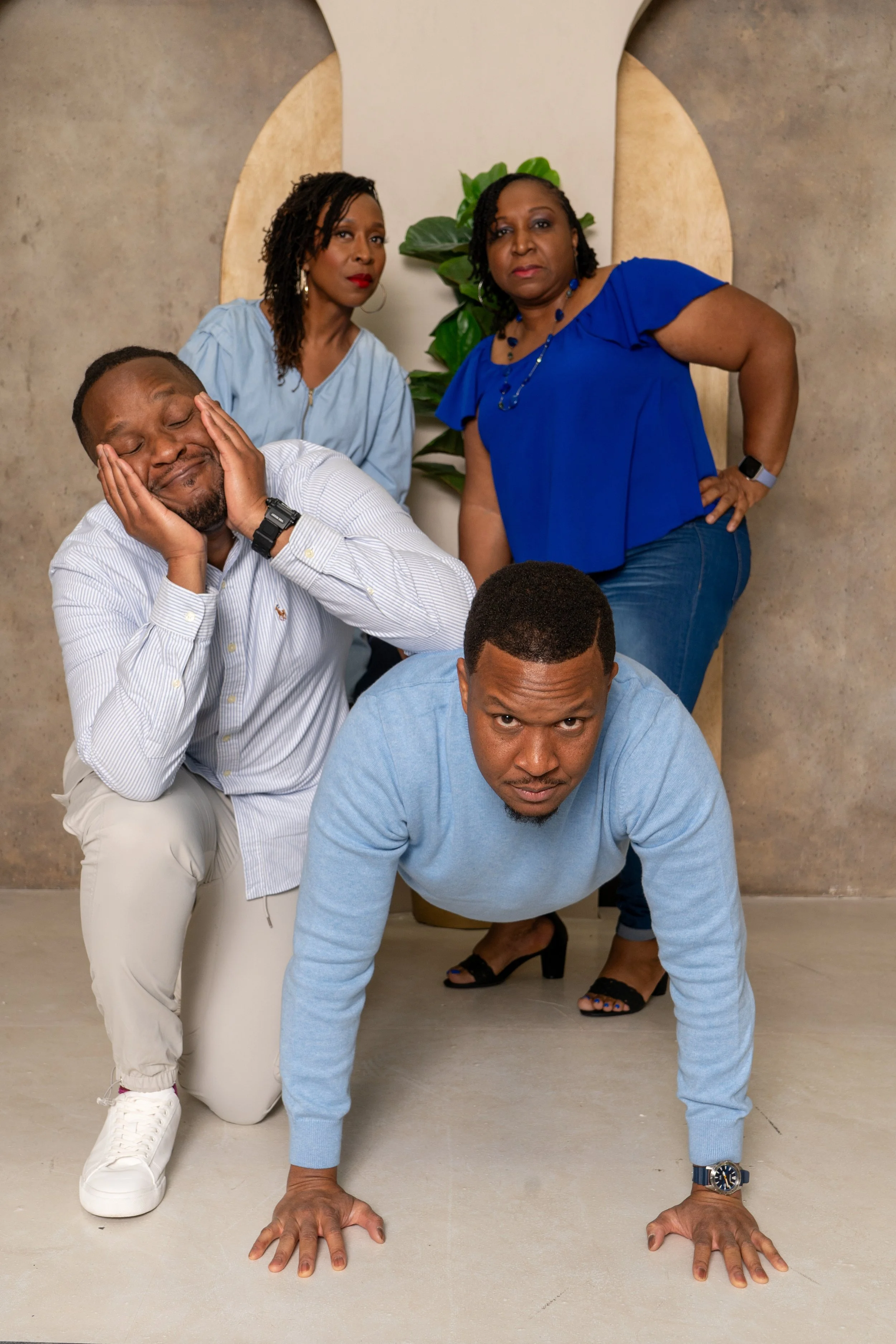 Four people posing in a studio with a neutral background, two women standing at the back, and two men crouching in front, one on hands and knees and the other on one knee, all making playful expressions.