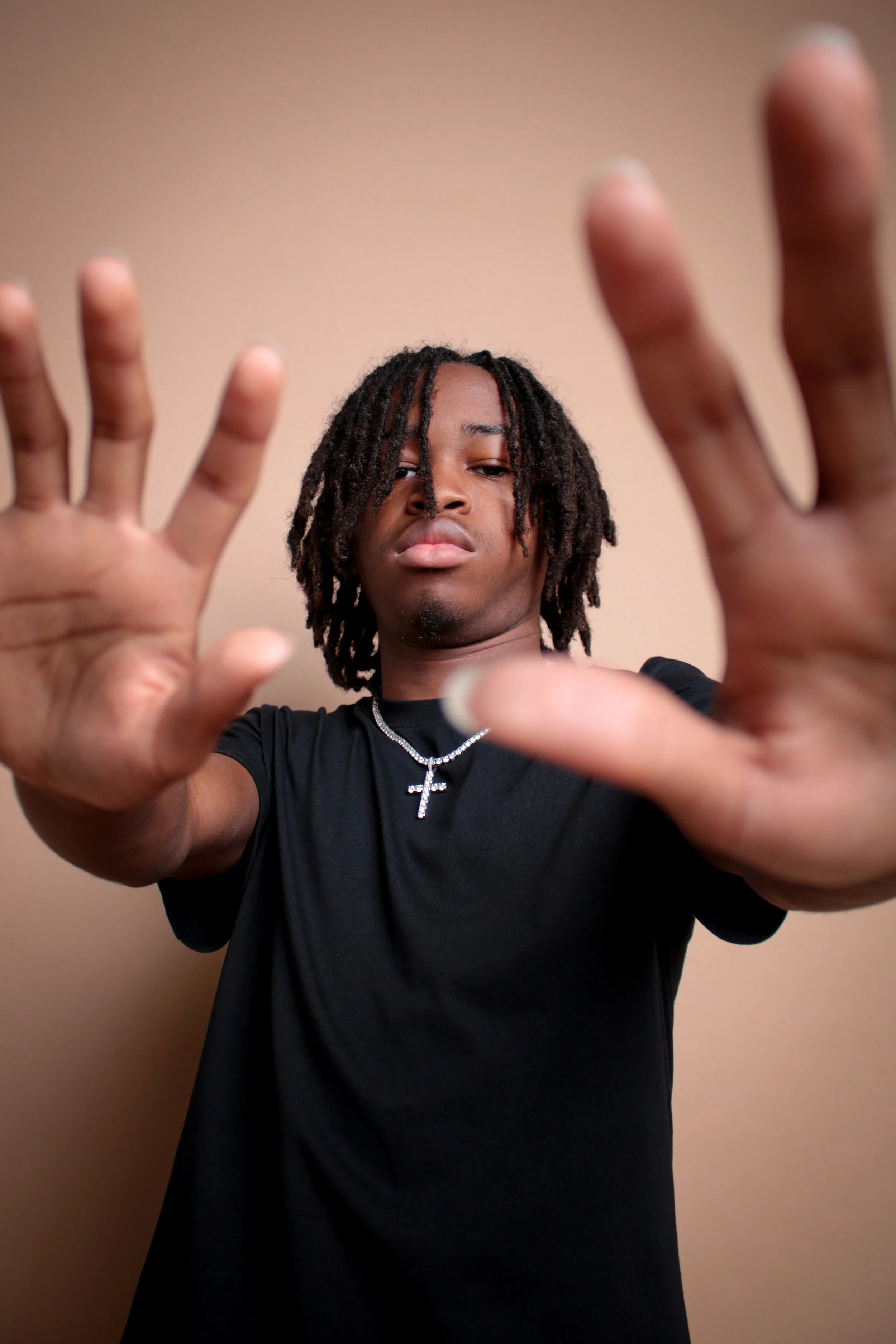 Young man with dreadlocks wearing a black t-shirt and a silver cross necklace, reaching towards the camera with both hands, against a plain beige background.