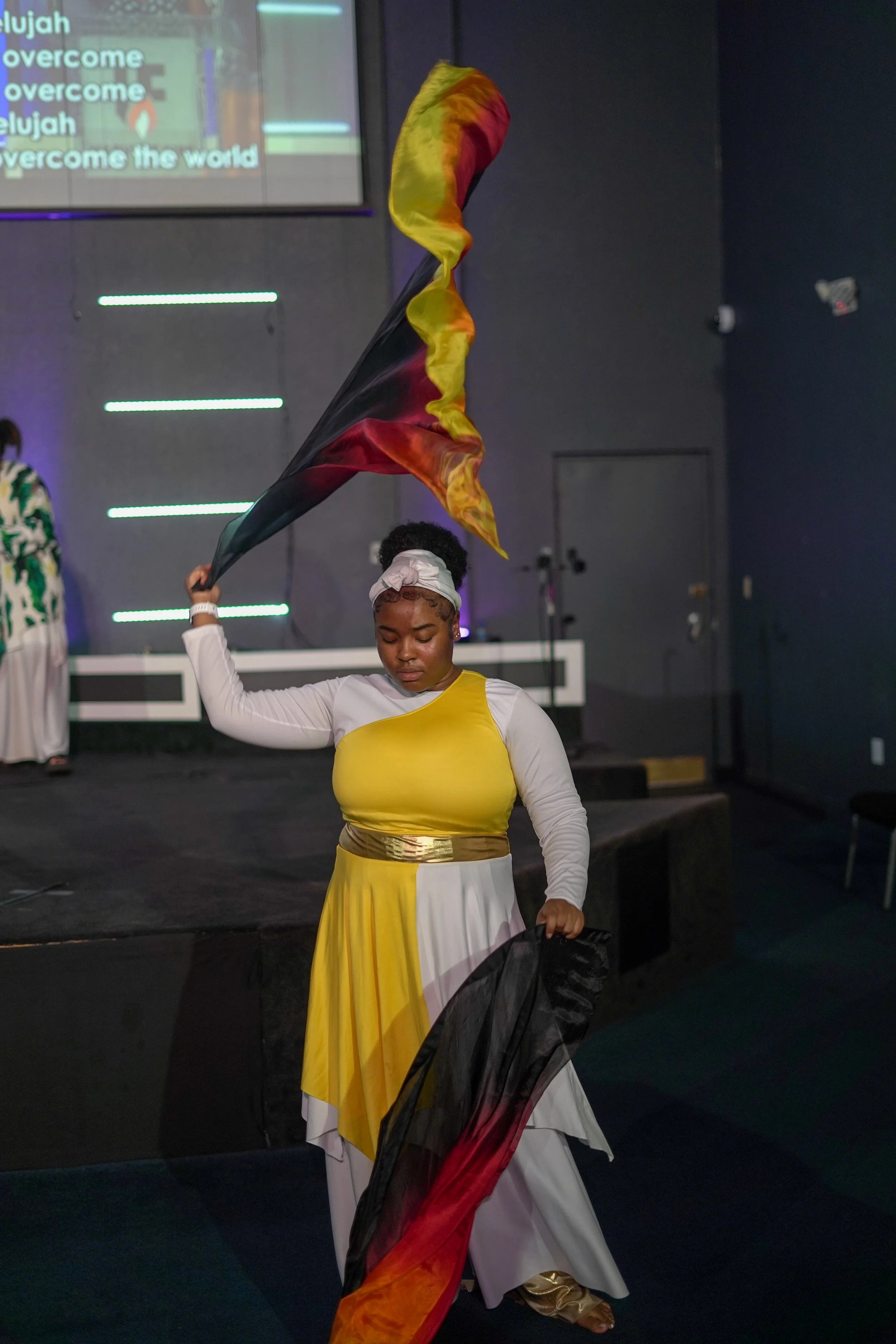 A woman dressed in a yellow and white dress is holding a multi-colored flag, standing on a stage in a church or performance space with a dark background and a large screen in the background displaying lyrics.