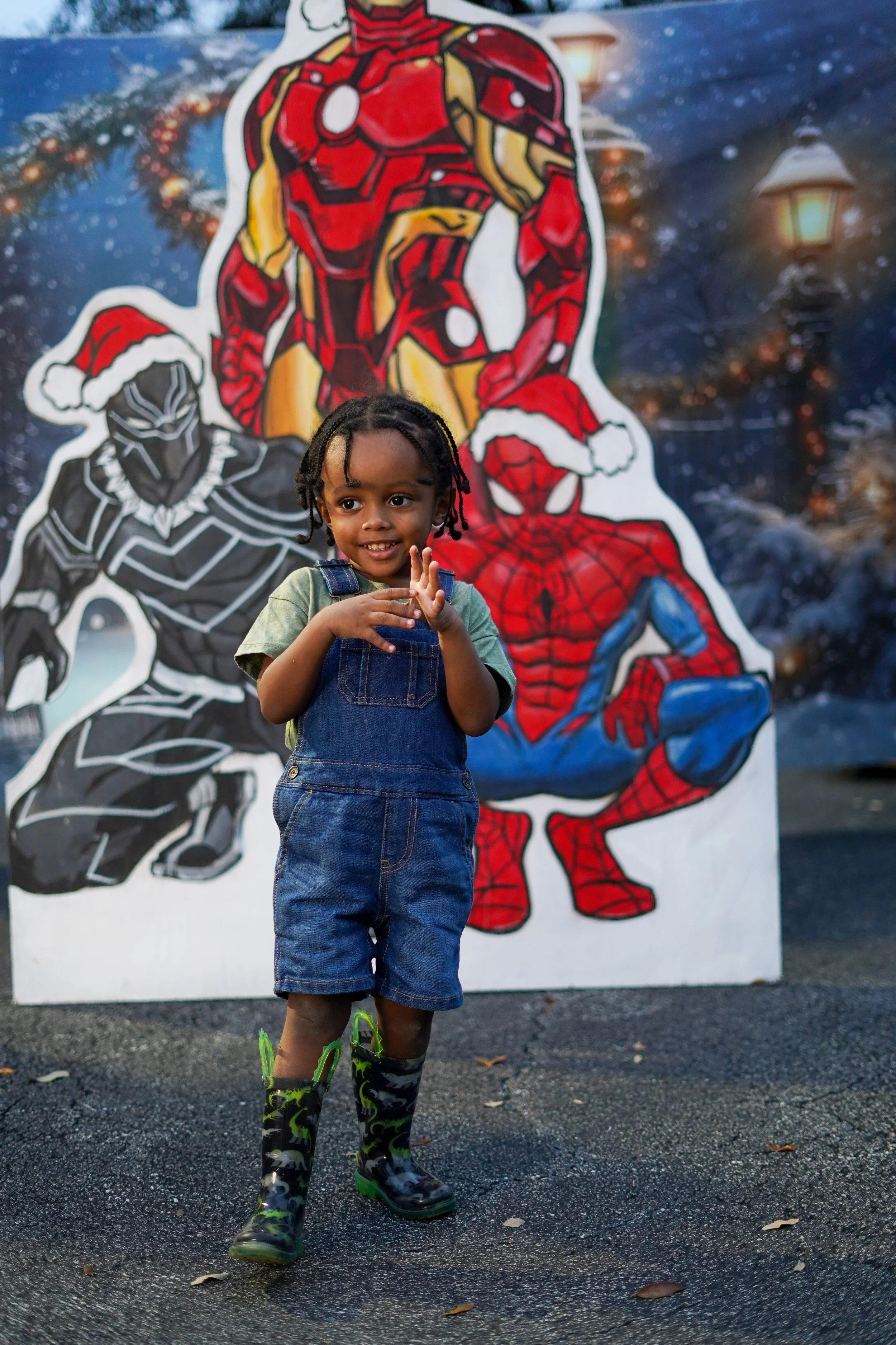 A young boy standing outdoors sporting a green shirt, denim overalls, and camouflage rain boots with a Spider-Man and Black Panther holiday decoration behind him.