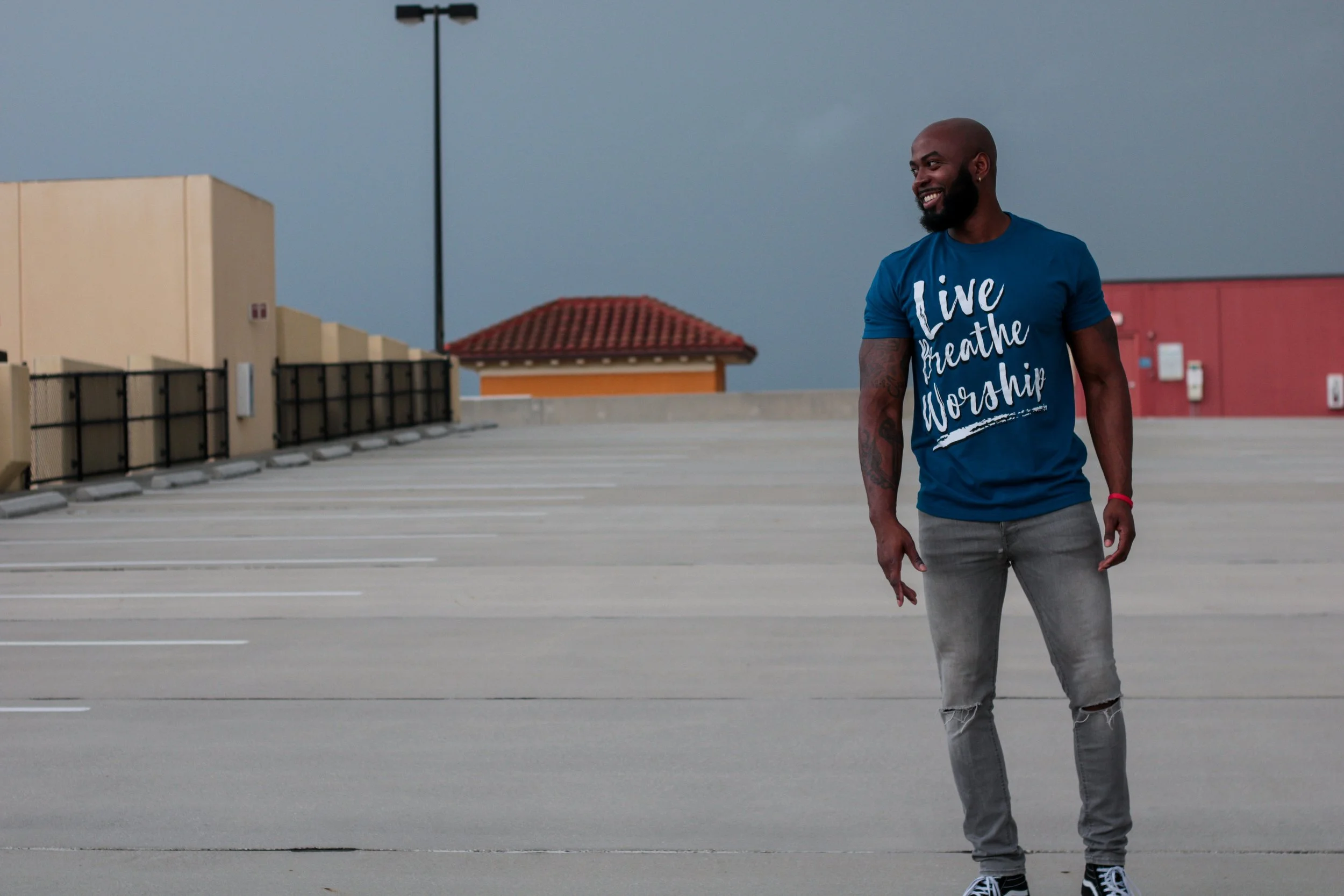 A smiling man wearing a blue T-shirt with the words 'Live Breathe Worship' standing in an empty parking lot during overcast weather.