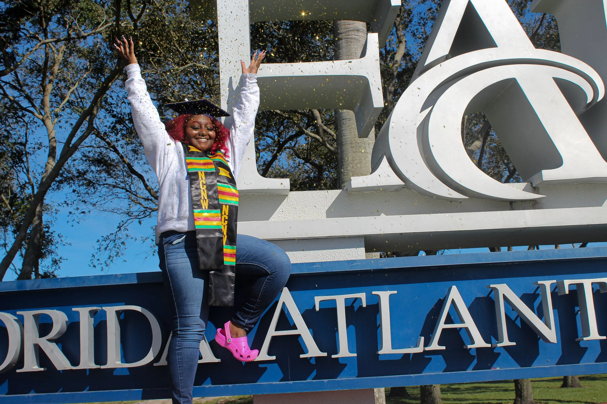 A young woman celebrating her graduation sitting on the 'Florida Atlantic' sign with arms raised, wearing a cap and gown, and a colorful stole, with a background of trees and a clear blue sky.