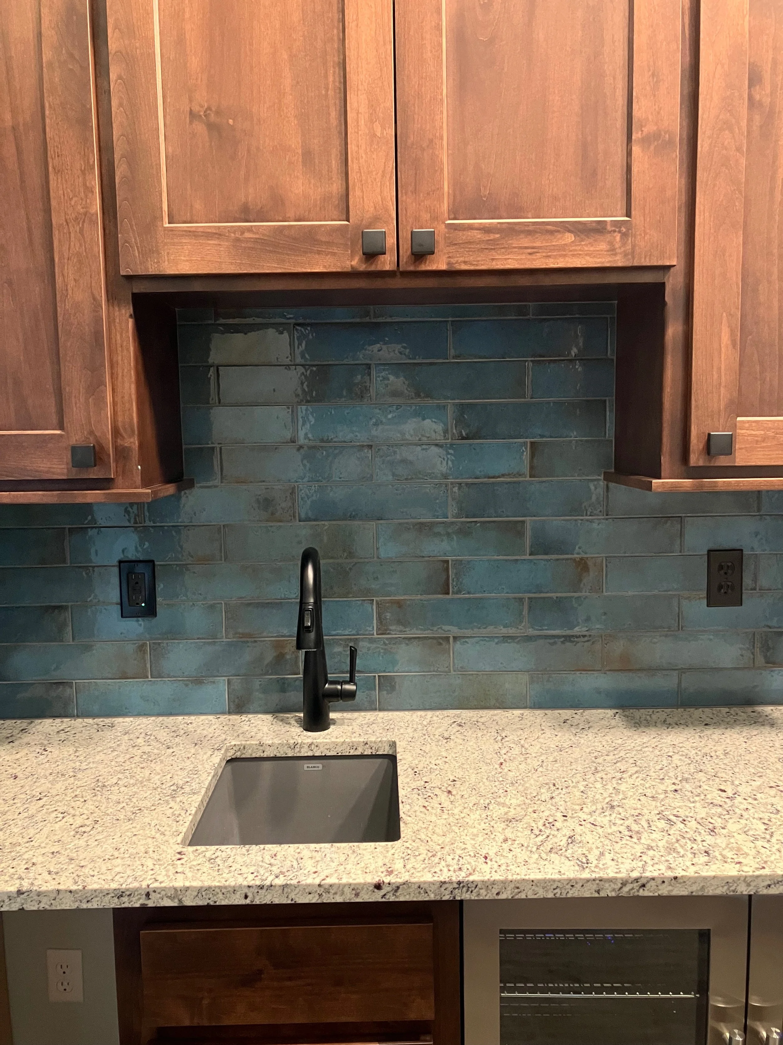 Kitchen sink with black faucet, granite countertop, blue brick backsplash, wooden cabinets, and electrical outlets.