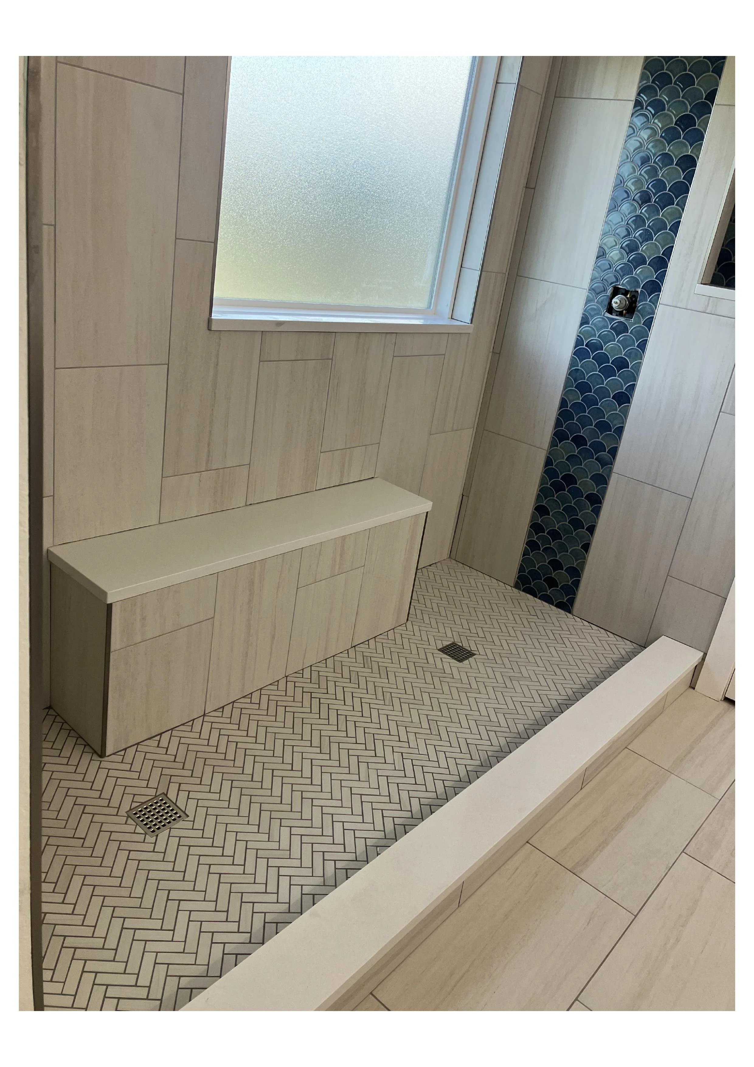 A modern bathroom shower area with beige tile walls, a frosted window, patterned floor tiles, and a decorative vertical strip of blue scalloped tiles.