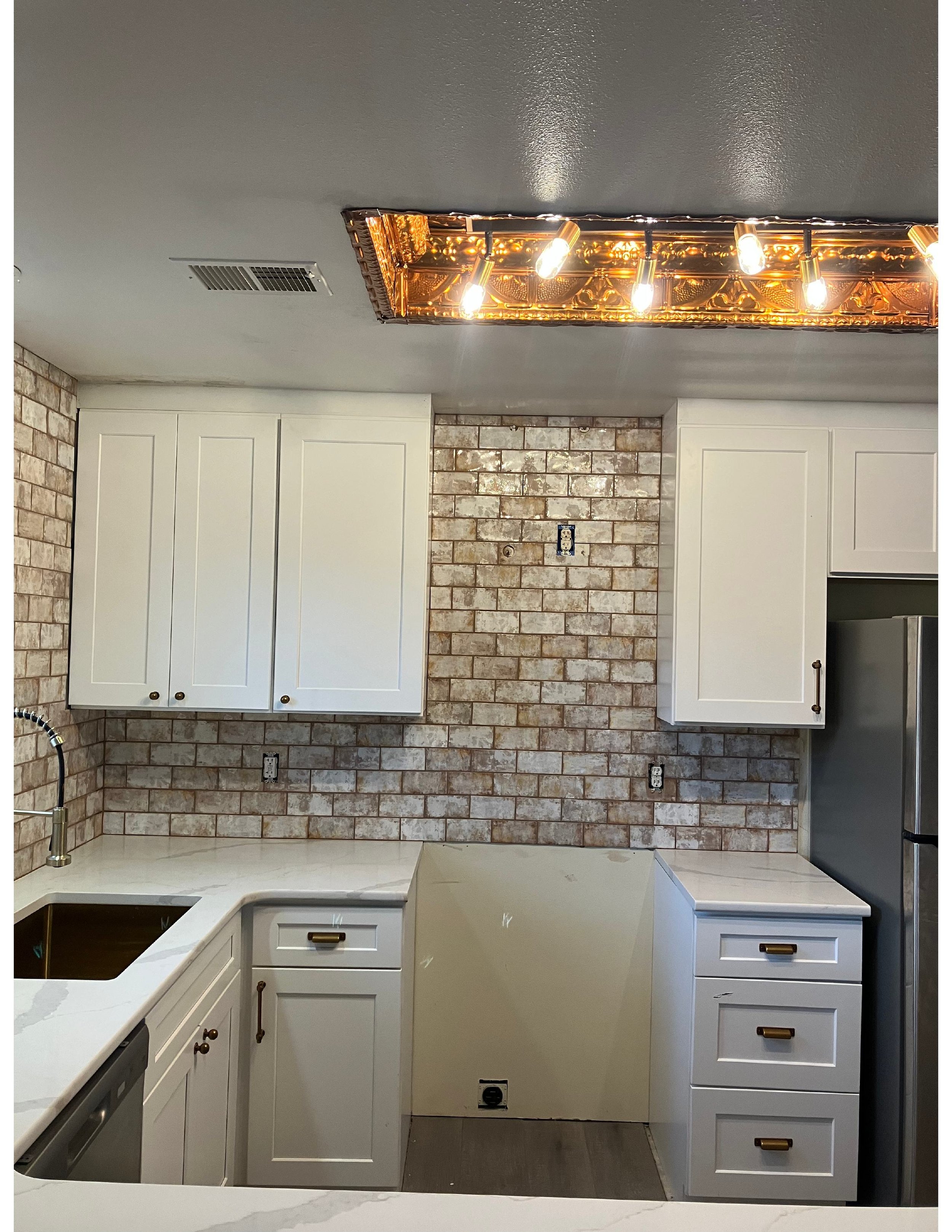 Kitchen with white cabinets, brick backsplash, and stainless steel refrigerator, under a decorative gold light fixture on the ceiling.