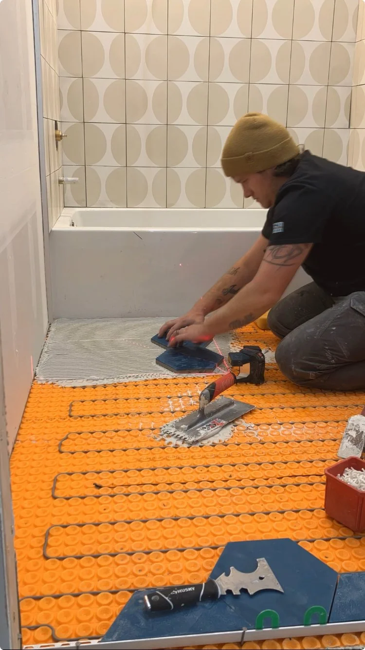 A person installing underfloor heating in a bathroom, kneeling on an orange foam mat with black heating cables, using tools and a laser measure.
