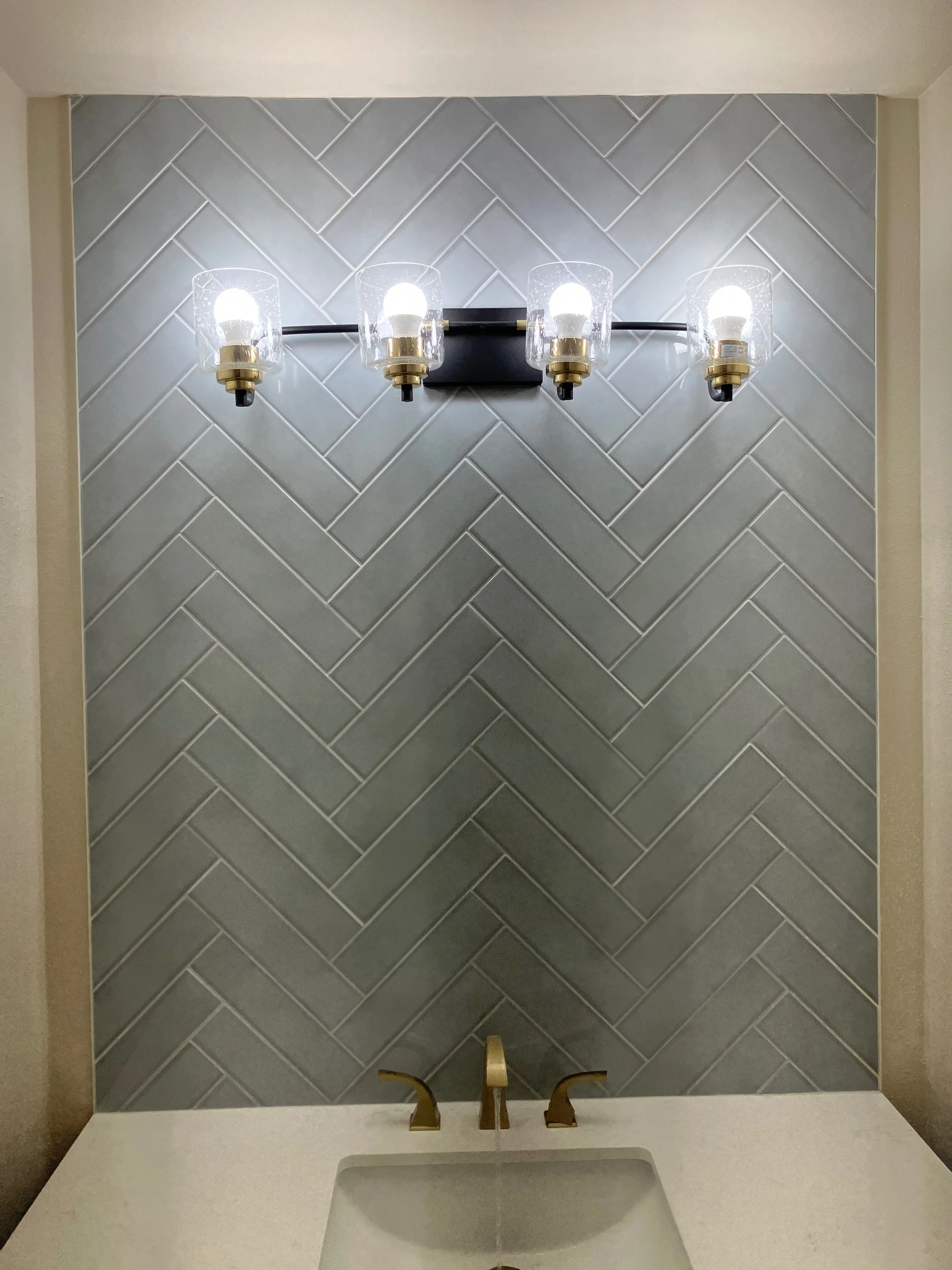 Bathroom sink with gold faucet, gray chevron tile wall, and four-wall sconce with glass shades.