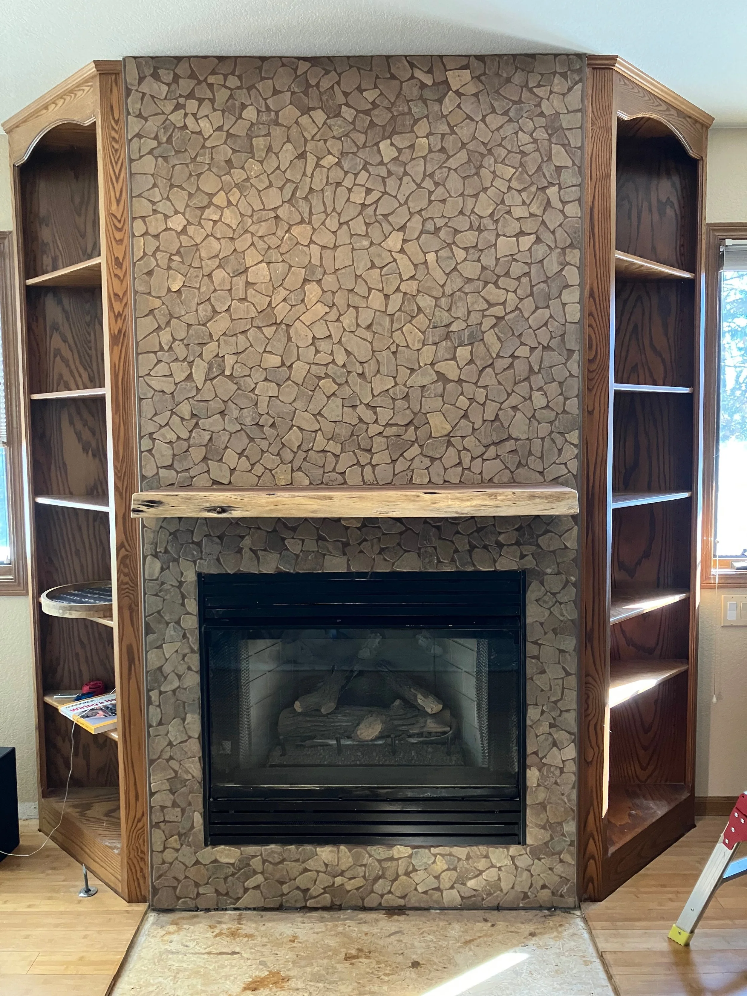Living room fireplace with a stone facade, surrounded by wooden built-in shelves and a wooden mantel, with a small window to the right and a light-colored rug on the floor.