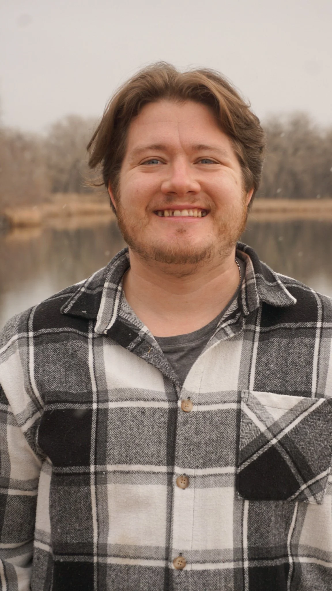 A smiling man with brown hair and a beard, wearing a black, white, and gray plaid shirt, standing outdoors near a body of water with trees in the background.