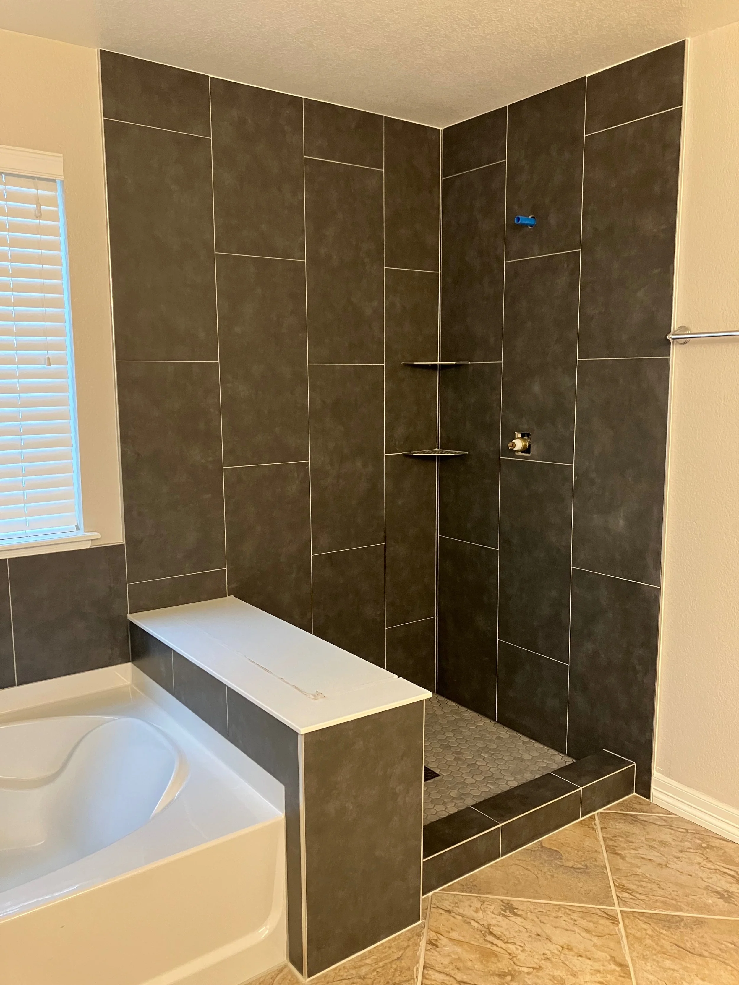 Bathroom with a bathtub and a walk-in shower area with dark gray tiles, a built-in corner shelf, a small blue pipe, and a valve, with beige tile flooring and a window with white blinds.