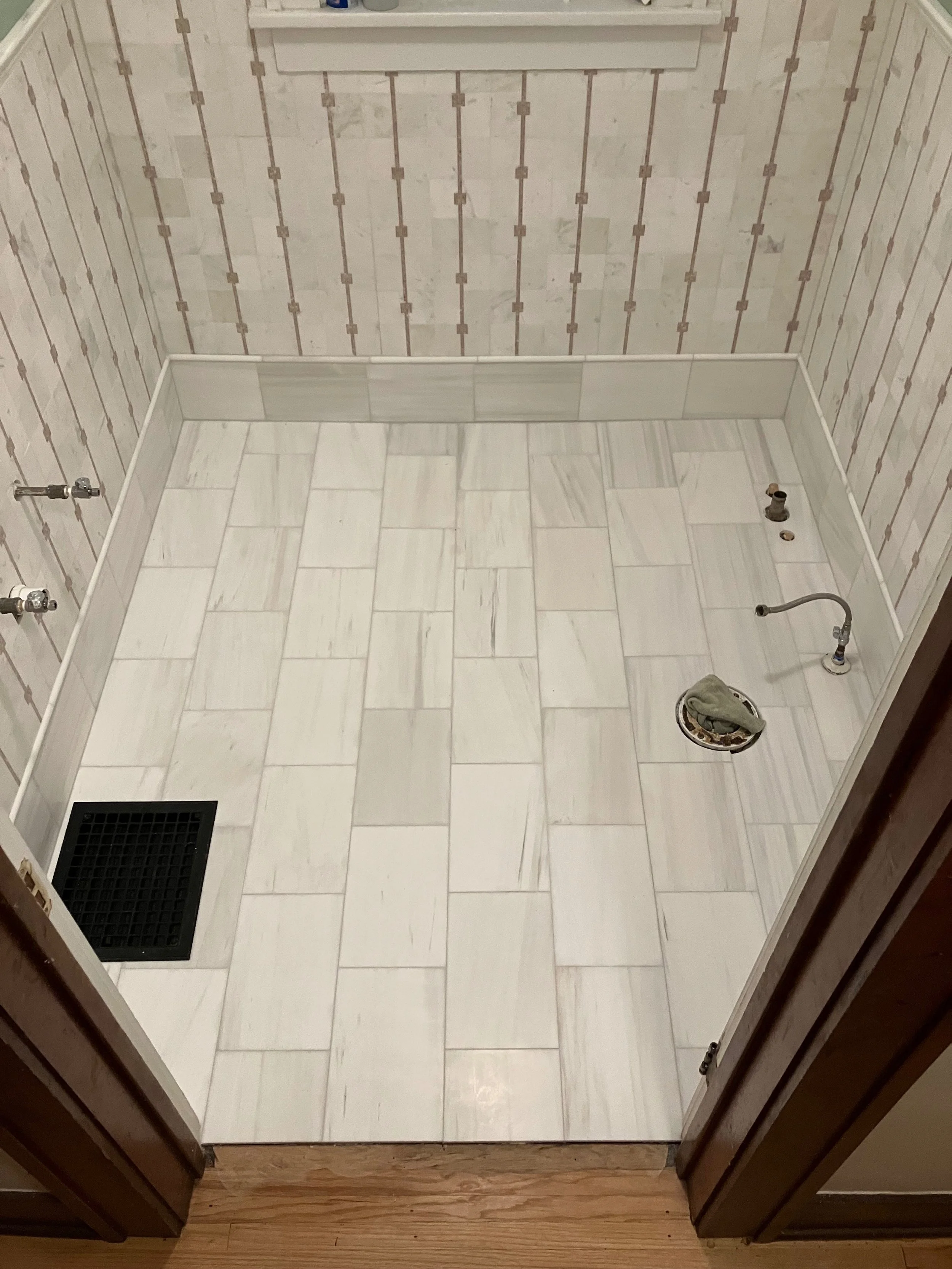 Empty bathroom shower area with white marble tiles on the floor, partially installed plumbing fixtures, and a black drain cover.
