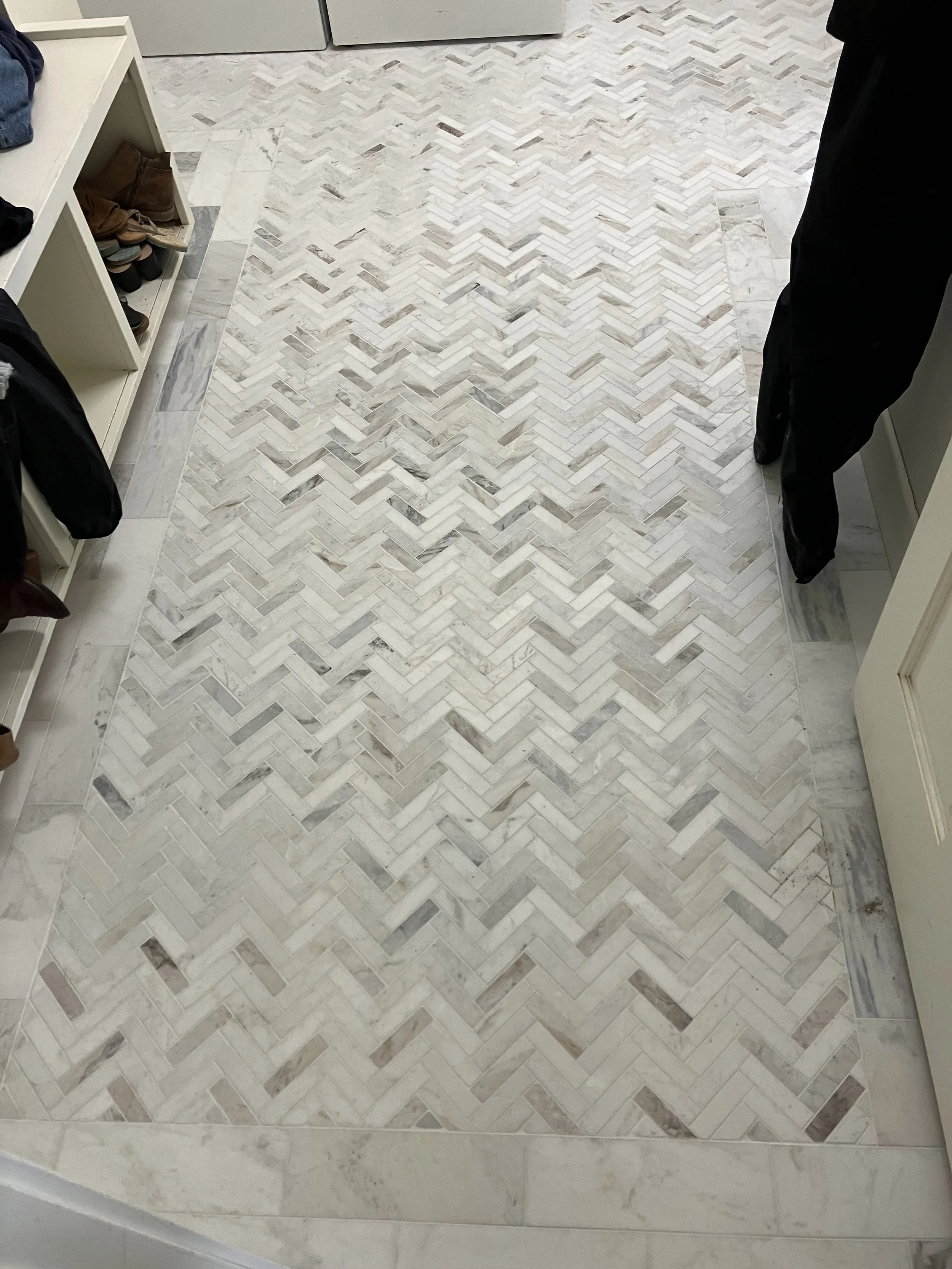 Close-up view of a herringbone marble tile floor with white, beige, and gray tones, in a room with white cabinetry and shelves containing shoes and clothing.