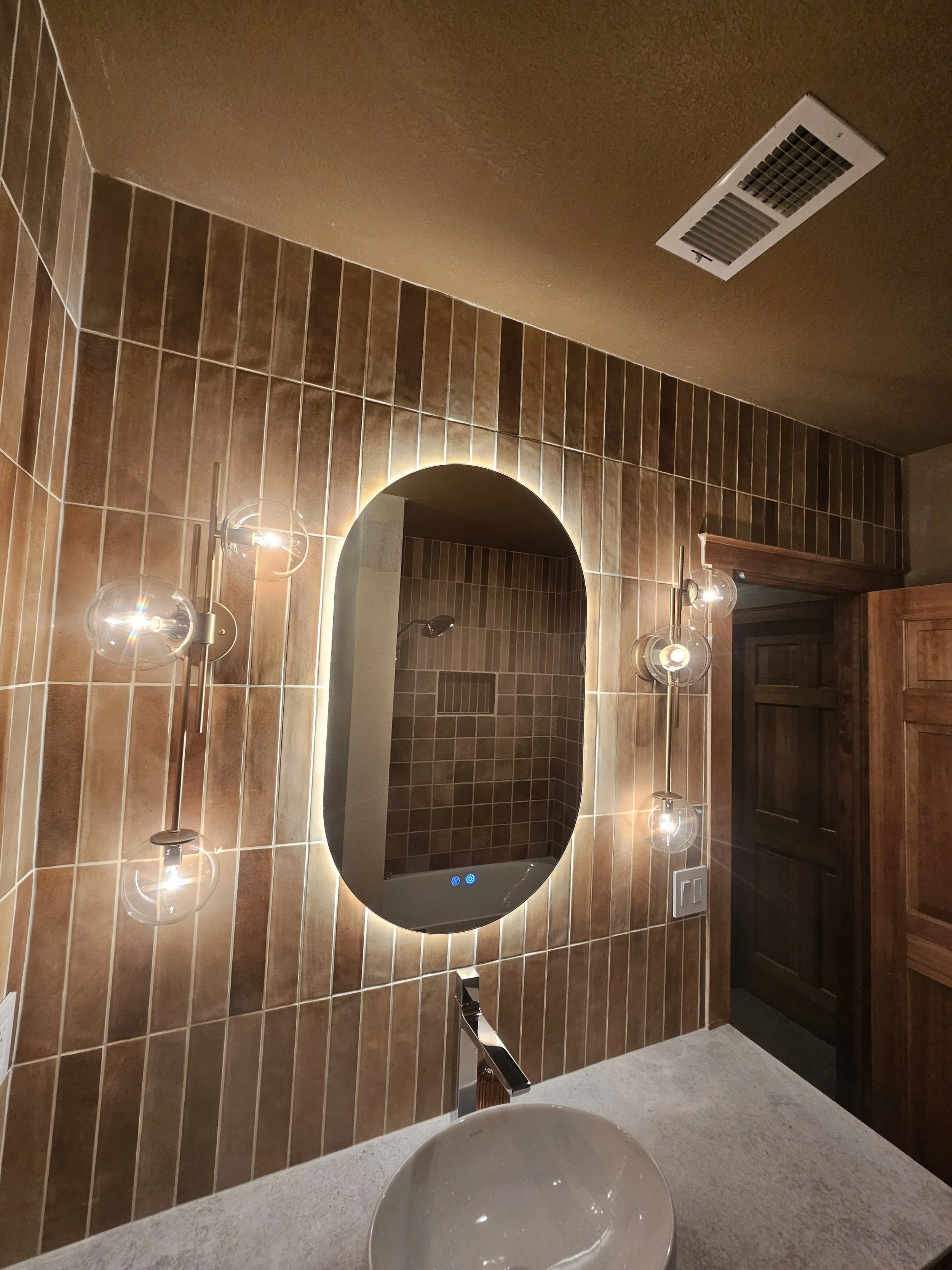 Bathroom with brown tiled wall, oval mirror with backlit LED lighting, modern wall sconces, wooden door, and a vessel sink on a light-colored countertop.