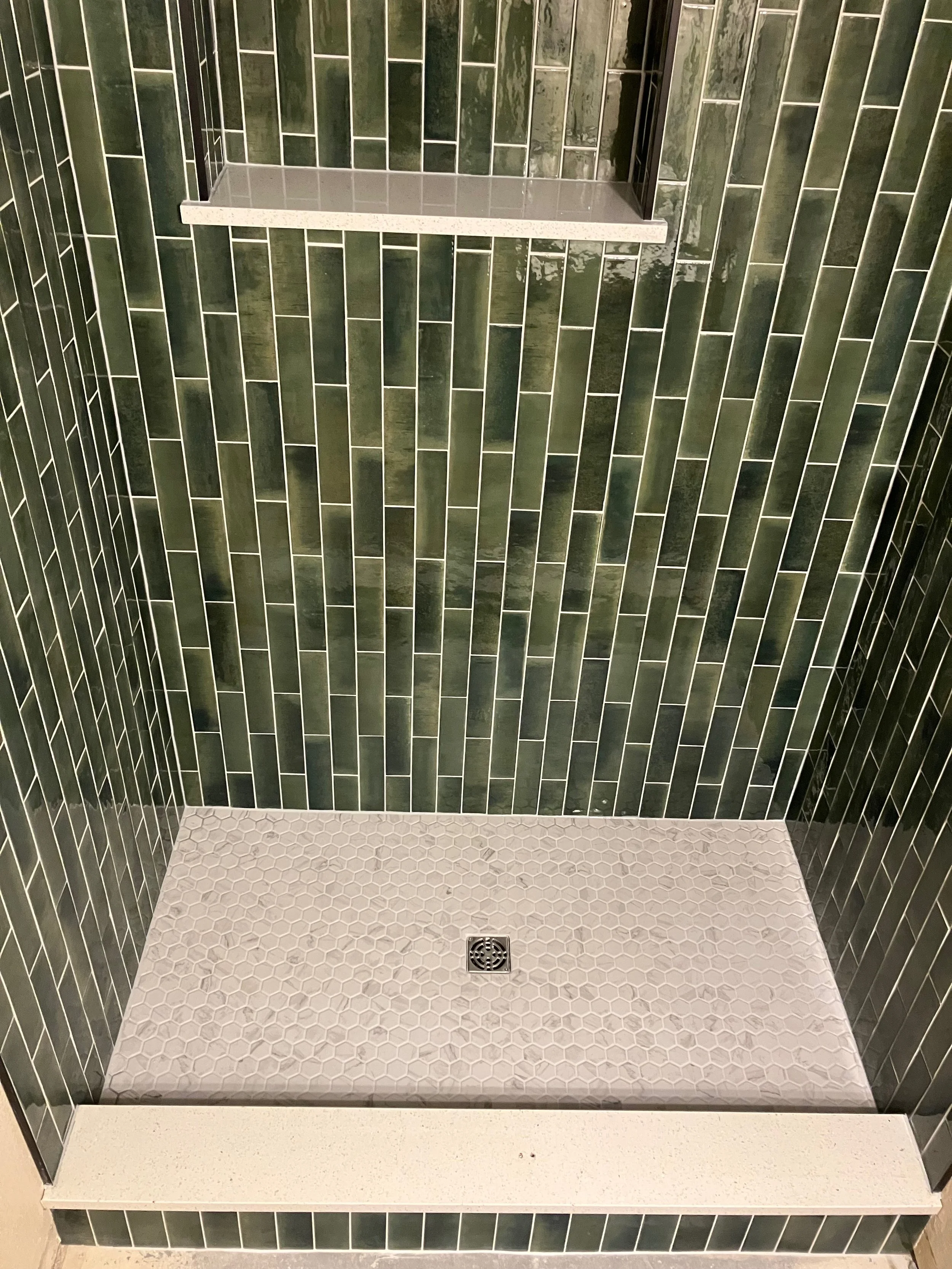 Green tiled shower with a small shelf and a metal drain on the white hexagonal tile floor.