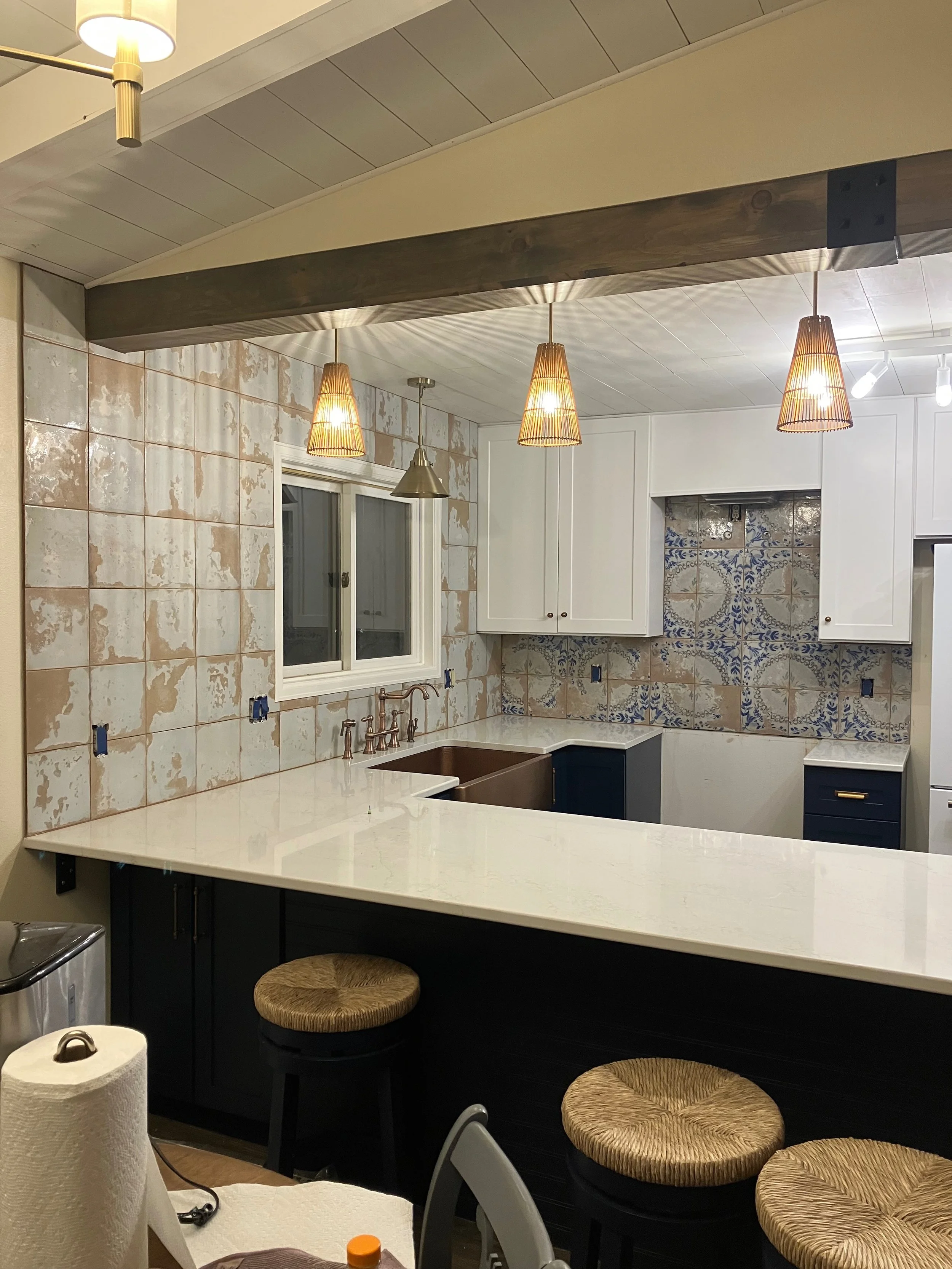Kitchen under renovation with white shiplap walls, a window, hanging pendant lights, a white countertop, and a dark island with bar stools.