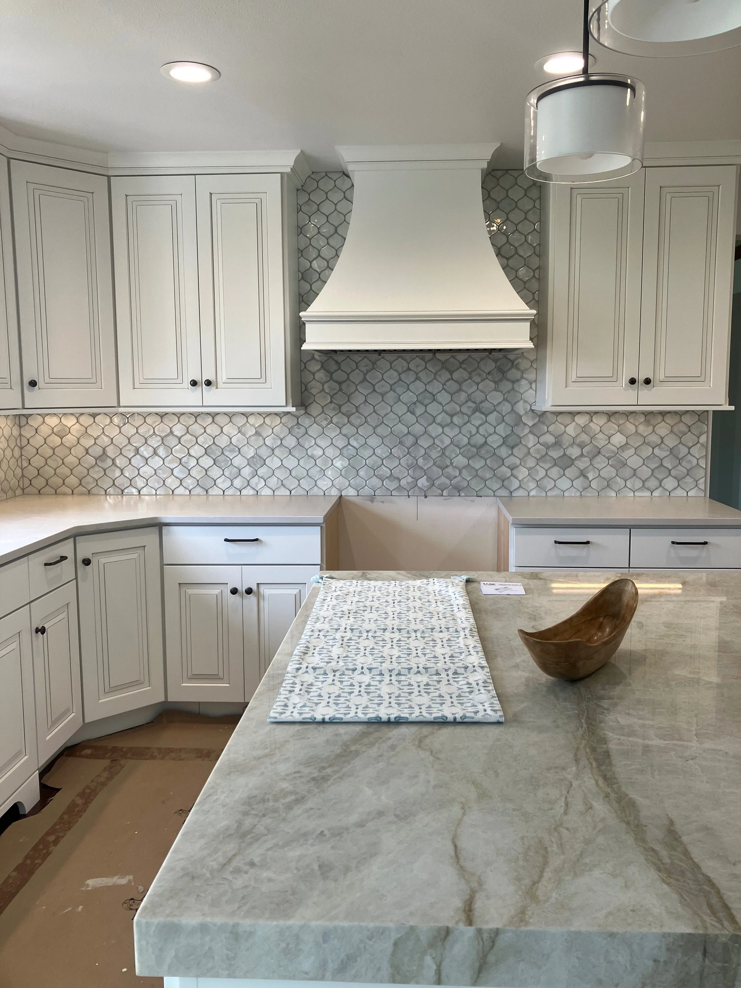 A modern kitchen with white cabinets, a decorative range hood, and a marble countertop island. There is a wooden bowl and a patterned cloth on the island. The backsplash features grey hexagonal tiles, and the ceiling has recessed lighting.