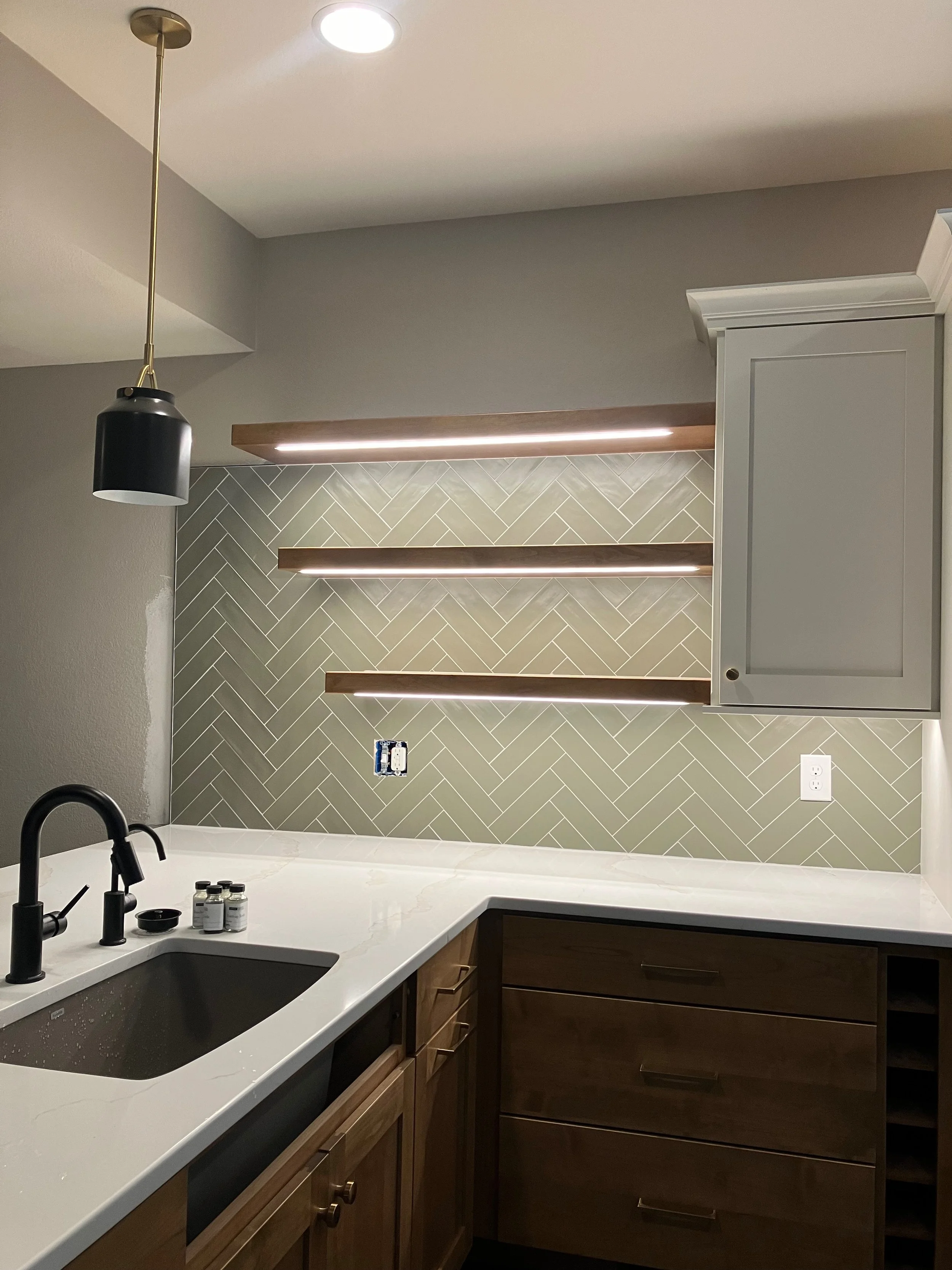 A modern kitchen with light gray cabinets, wooden drawers, white marble countertop, black sink and faucet, three small bottles, and a black pendant light. The accent wall features gray herringbone tiles with three wooden shelves and integrated lighti