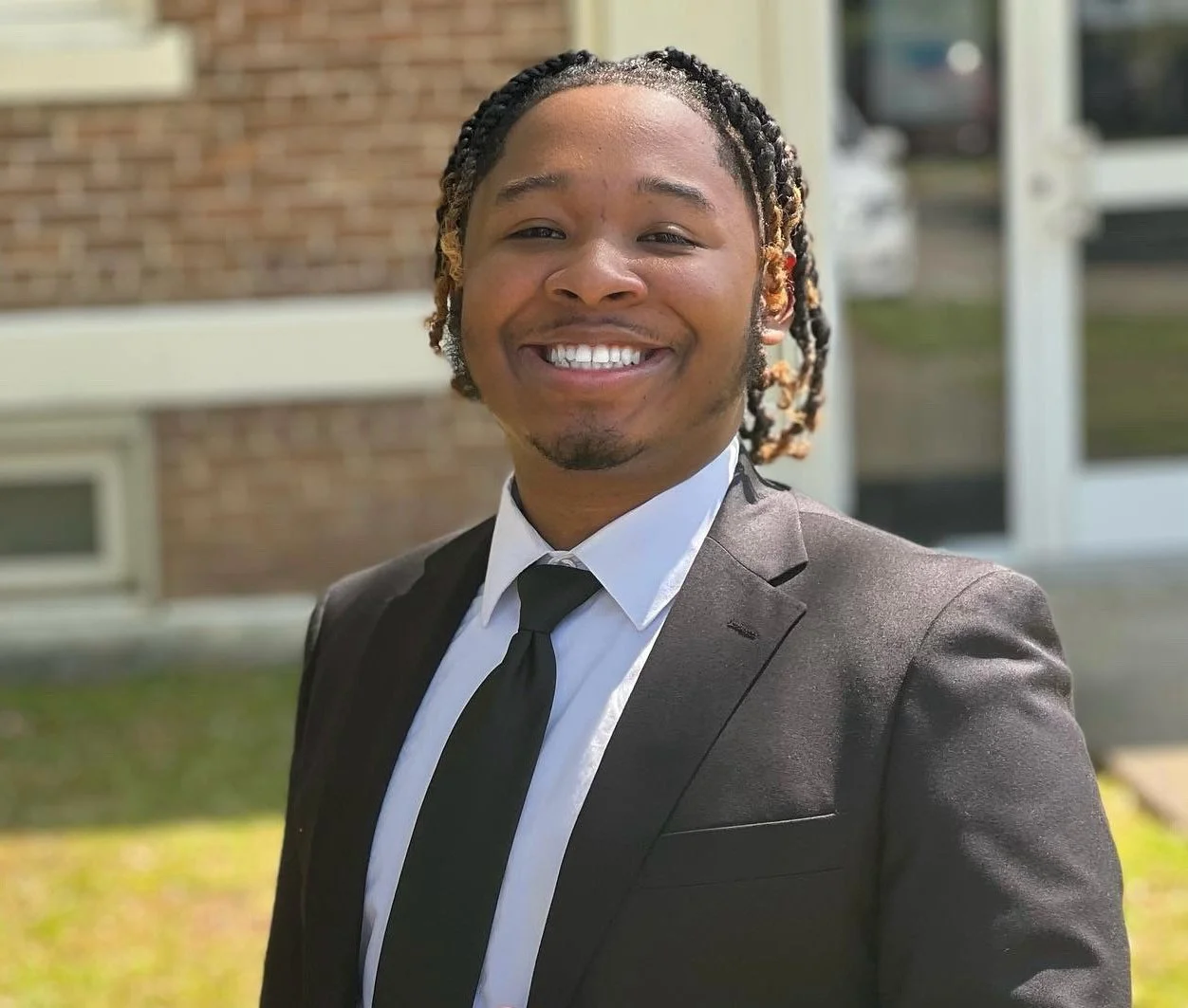 A smiling man in a black suit, white shirt, and black tie standing outdoors in front of a brick building.