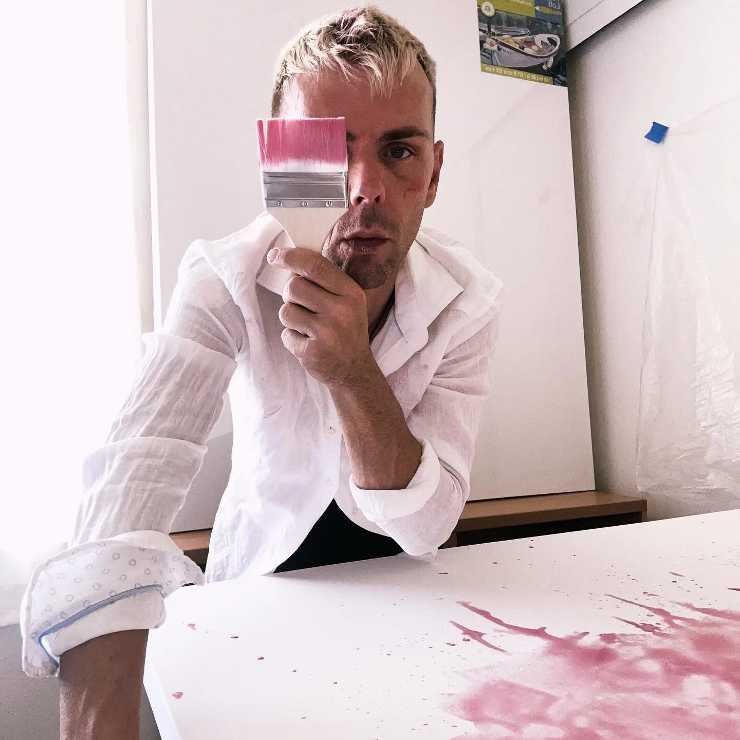 A man with blonde hair holding a paintbrush, looking at the camera with a serious expression, with pink paint on the table and a white shirt with rolled-up sleeves.