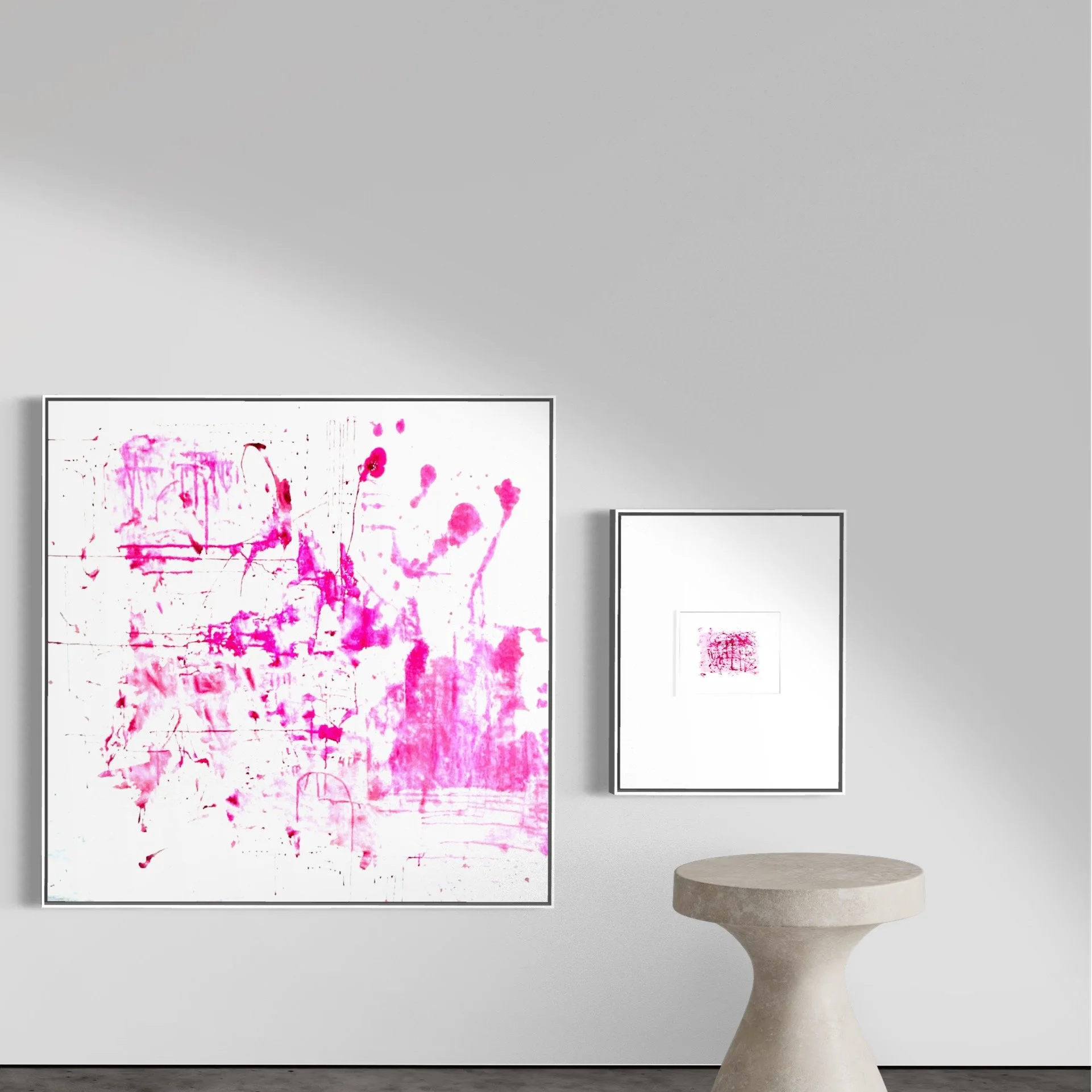 Two framed abstract art pieces with prickly pear pink paint splashes on a white gallery wall, and a small round concrete table in front.